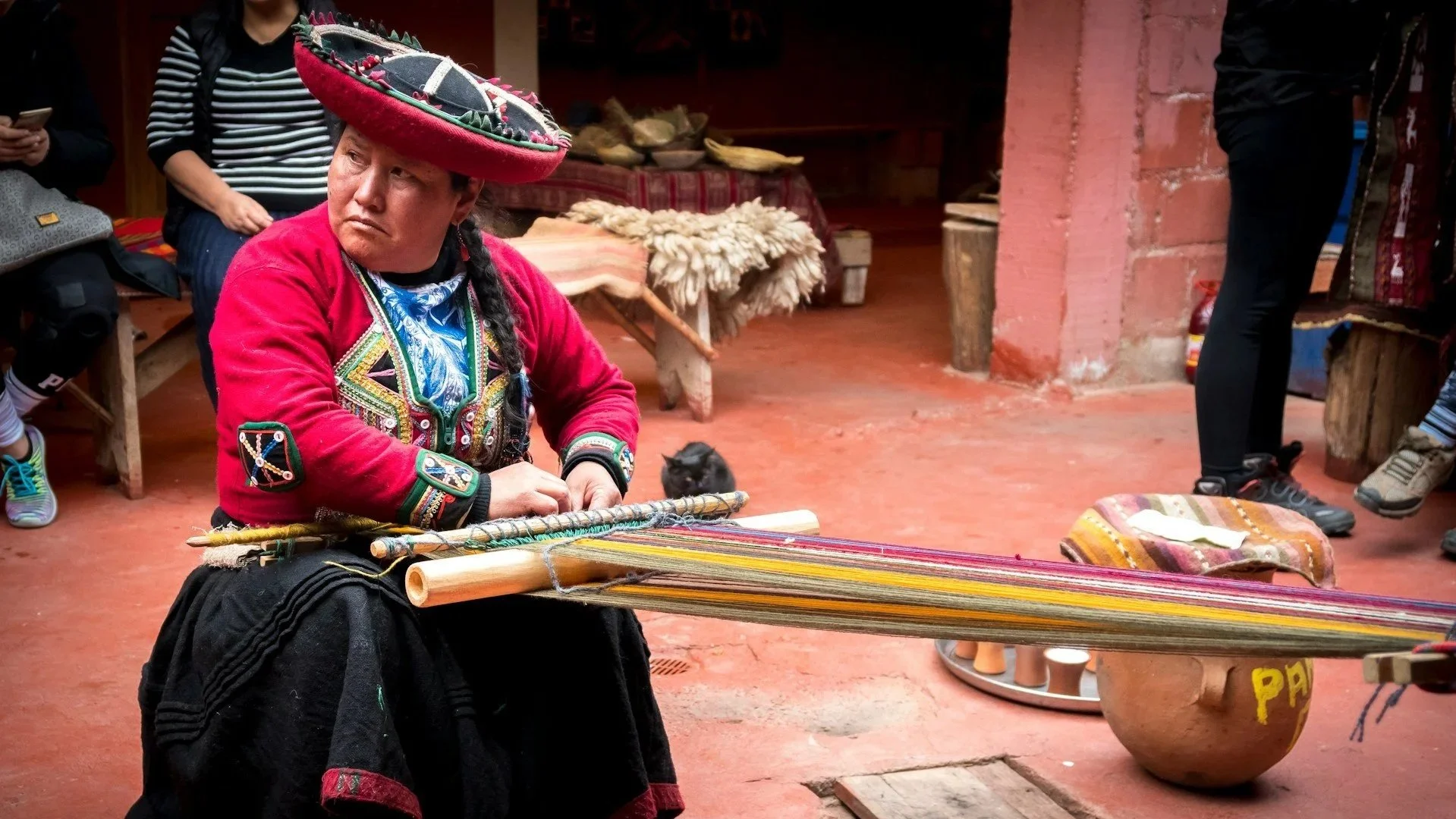 An Andean woman in vibrant traditional dress weaving colorful textiles on a backstrap loom.