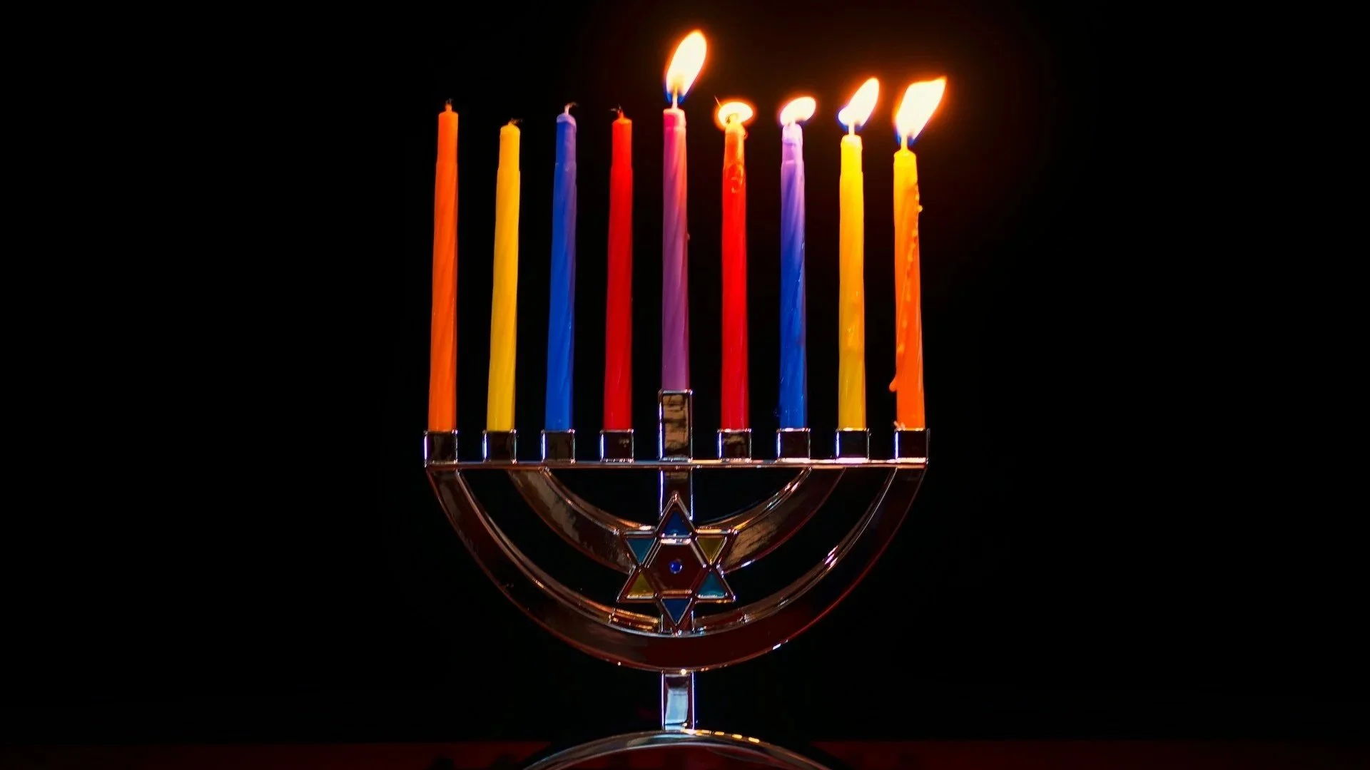 A silver Hanukkah menorah with nine colorful candles, five of which are lit, glowing against a black background.