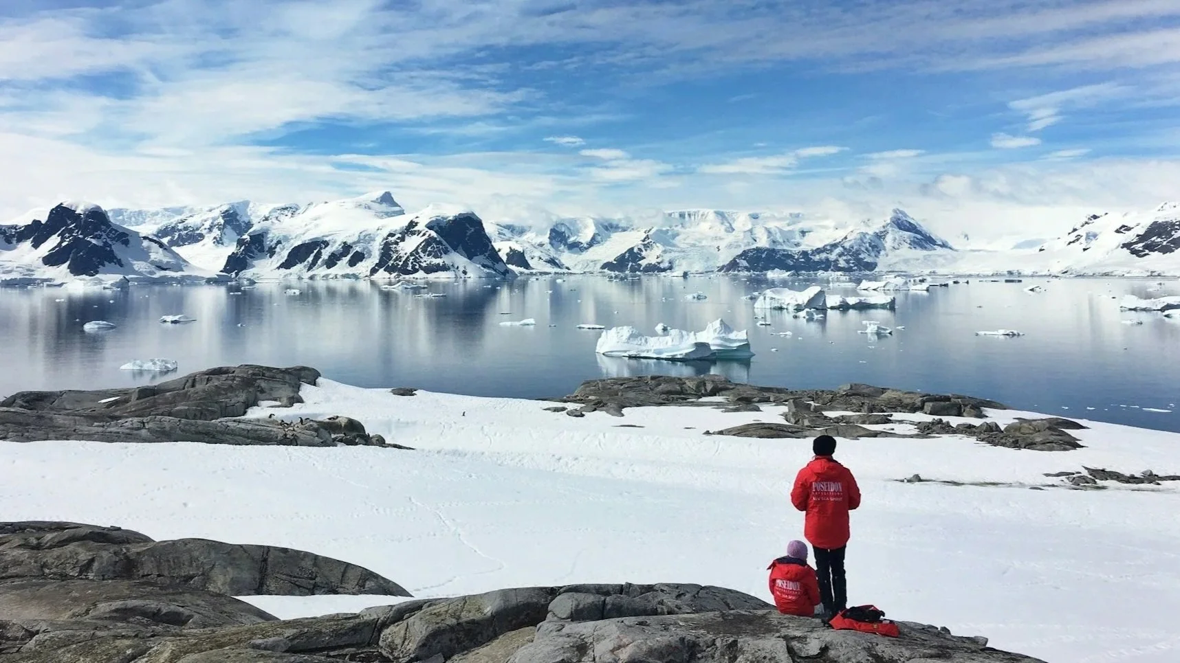 Two people in red parkas standing on a snowy rocky outcrop looking out over a calm sea filled with icebergs and surrounded by snow-capped mountains.