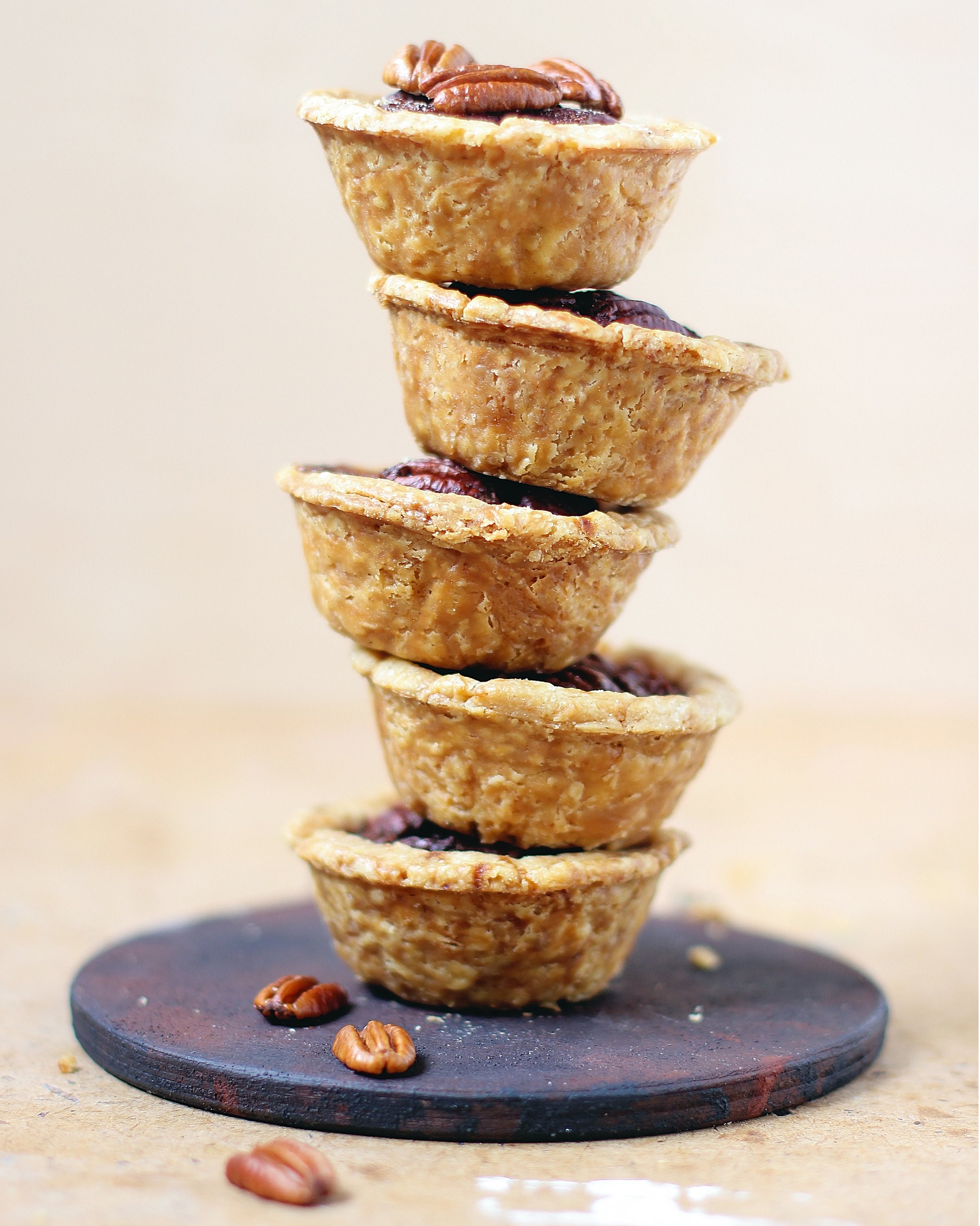 A vertical stack of five mini butter tarts topped with whole pecans sitting on a dark wooden coaster.