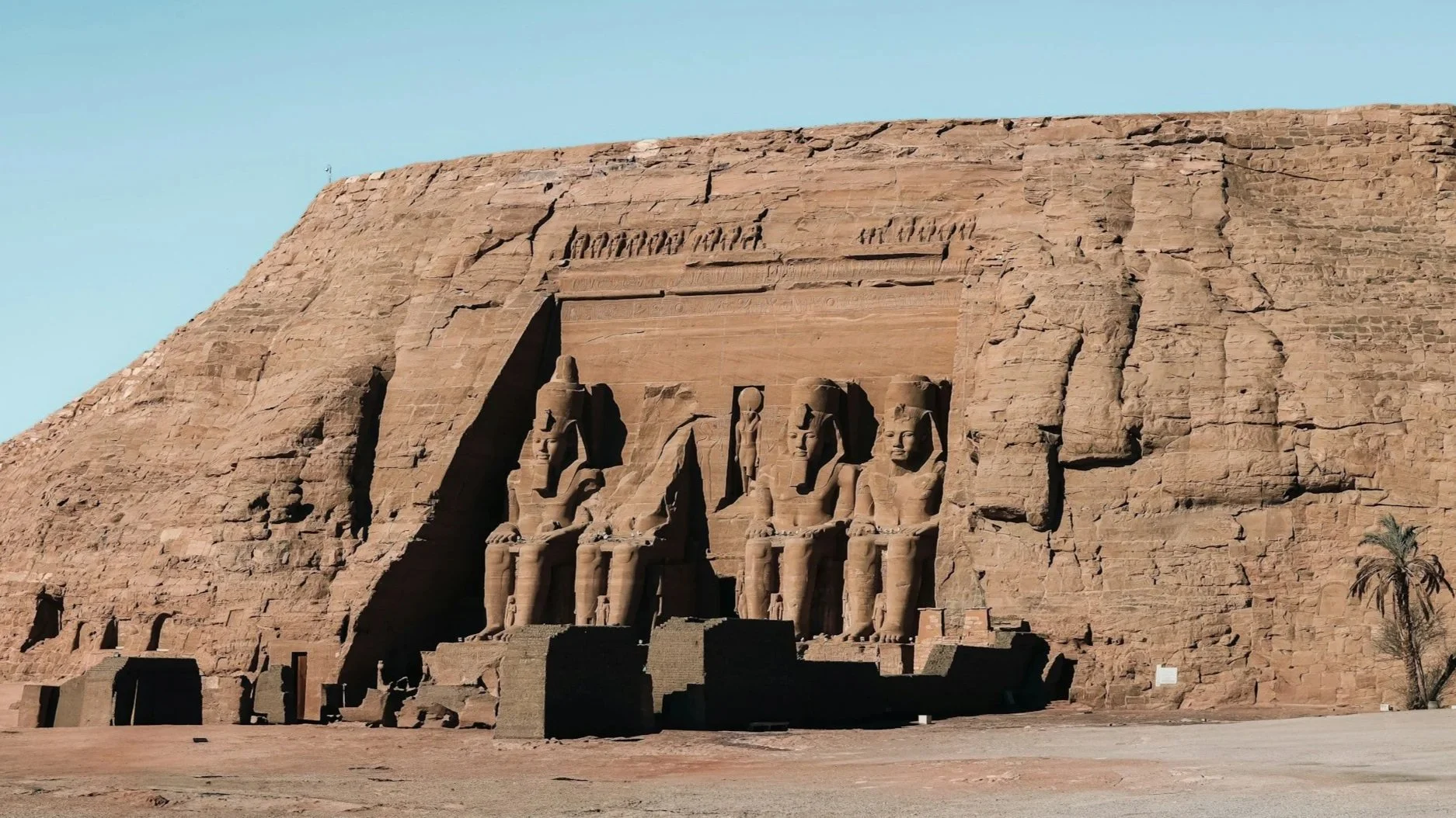 The Great Temple of Abu Simbel featuring four massive seated statues of Ramesses II carved into a sandstone cliff under a clear blue sky.