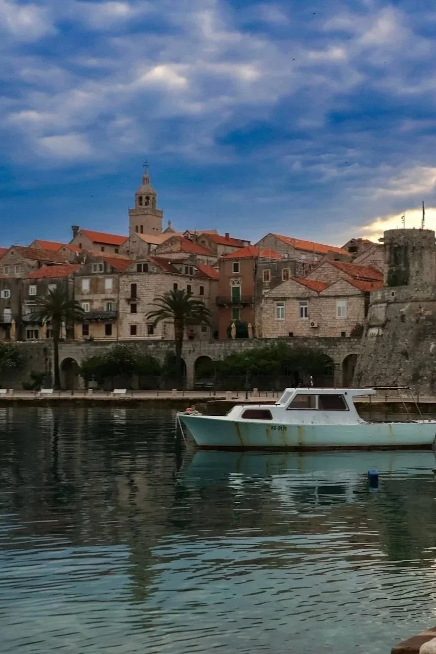 A small boat floats in a calm harbor at dusk, with the fortified stone walls and red-roofed buildings of Korčula Town in the background.