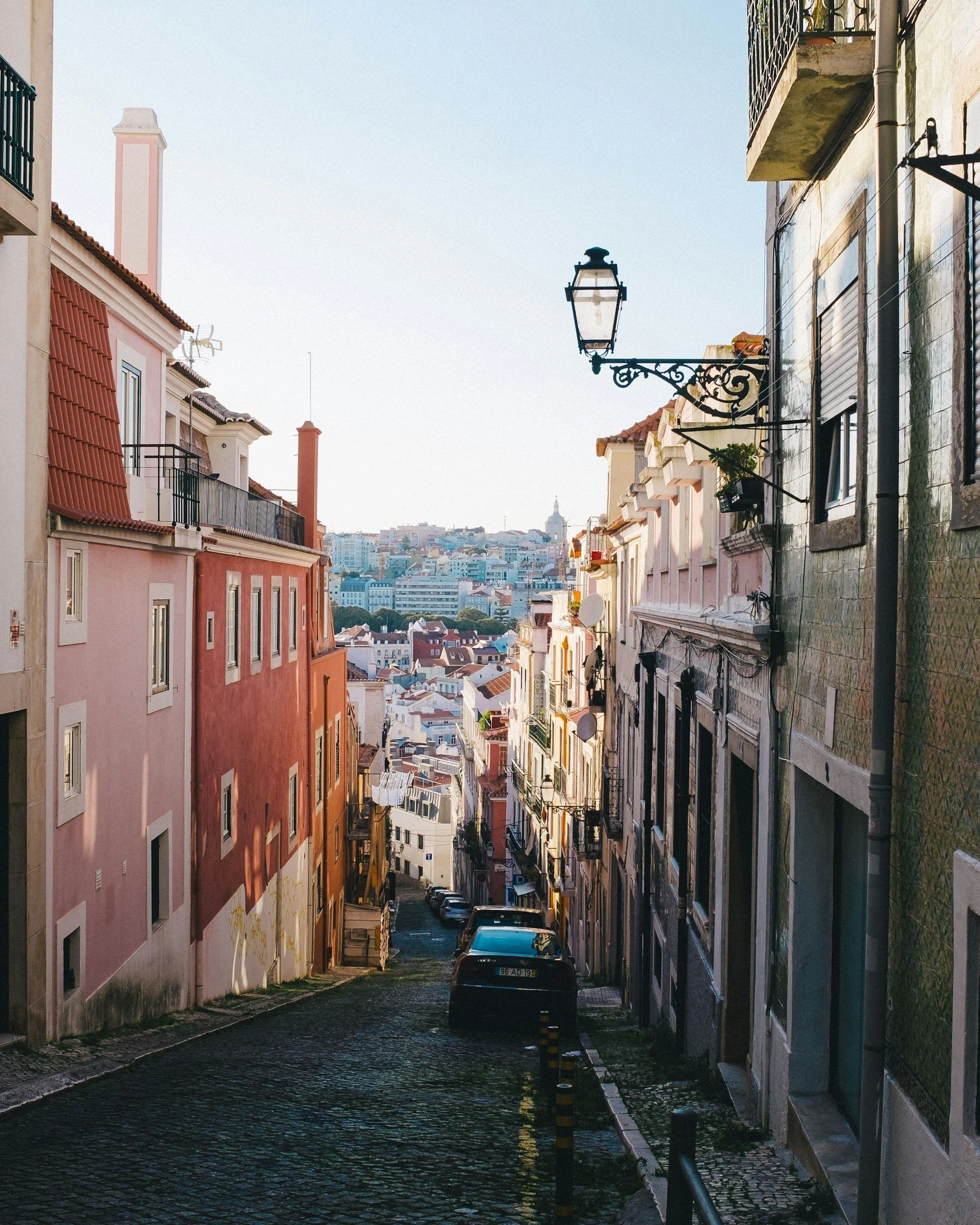 A narrow, sunlit cobblestone street in Lisbon, Portugal, lined with colorful pastel buildings and parked cars leading toward a city view.