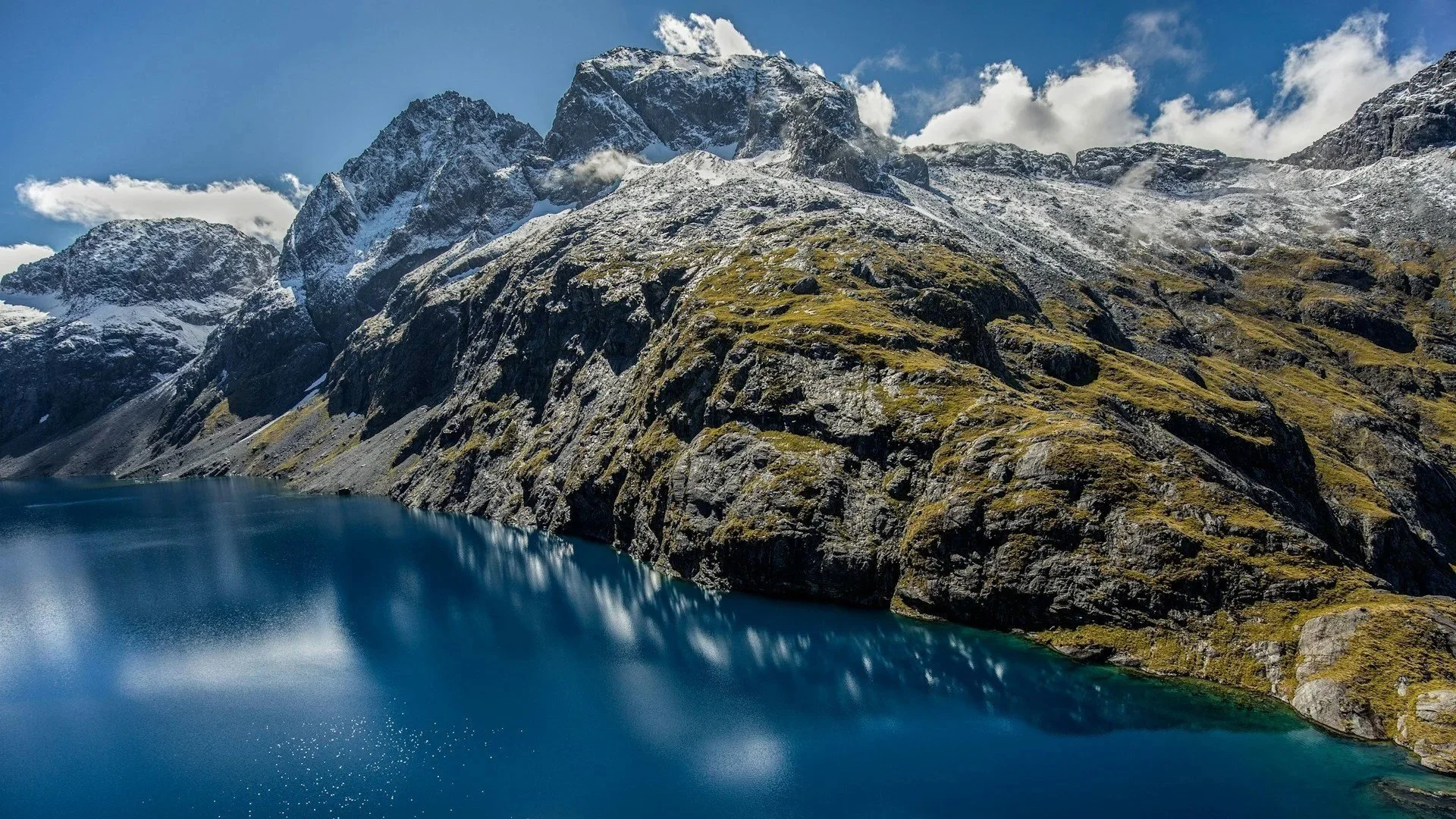 High-angle shot of a deep blue alpine lake surrounded by steep, snow-dusted mountains.