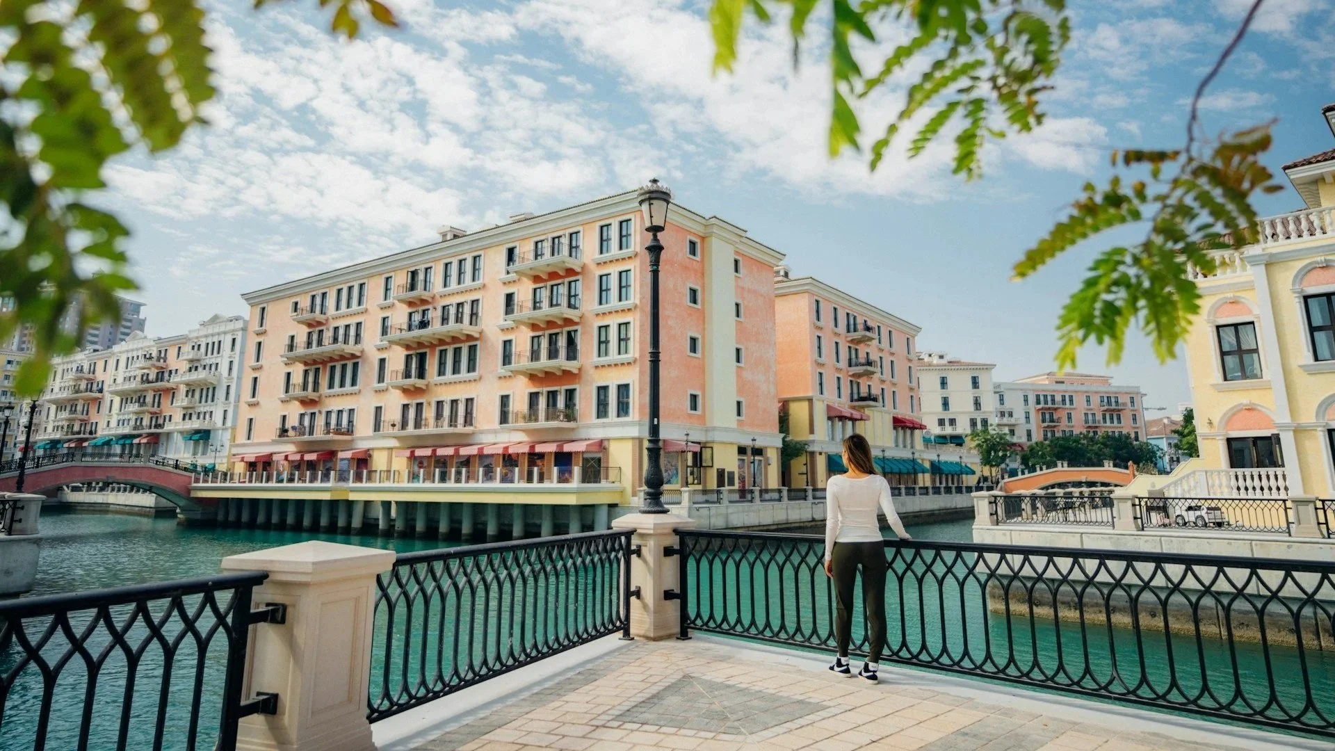 A woman stands at a railing looking across a turquoise canal at Mediterranean-style colorful buildings at The Pearl-Qatar.