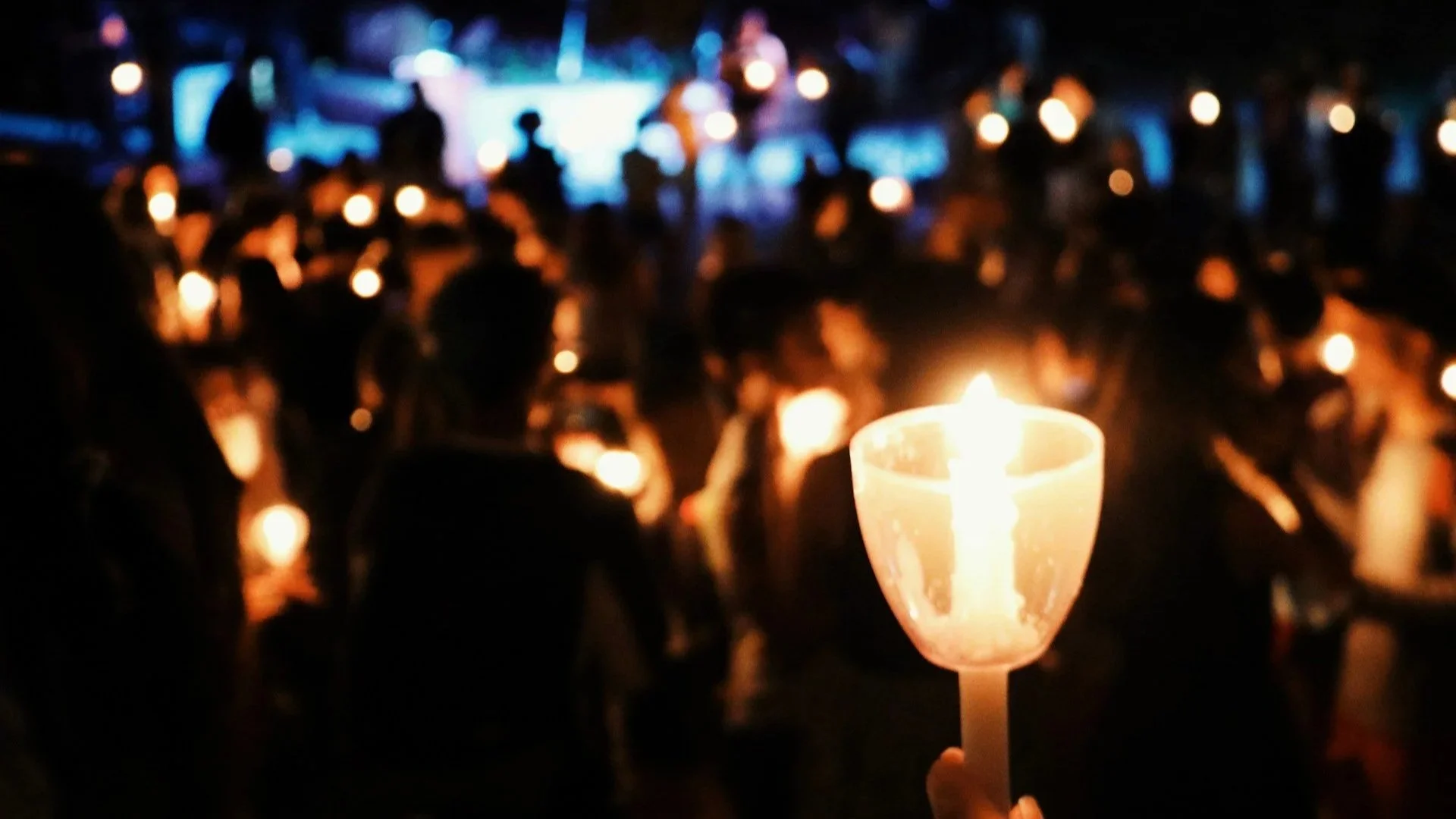 A close-up of a hand holding a lit candle in a clear plastic cup at a nighttime vigil, with a blurred crowd of people and glowing candles in the background.