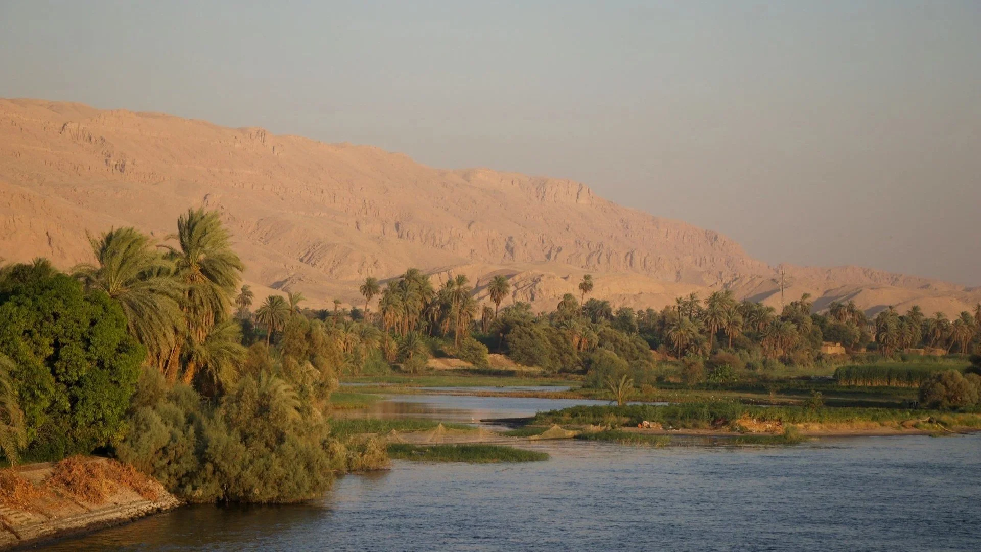 A peaceful view of the Nile River at sunset, lined with lush green palm trees and set against rugged, desert mountains.
