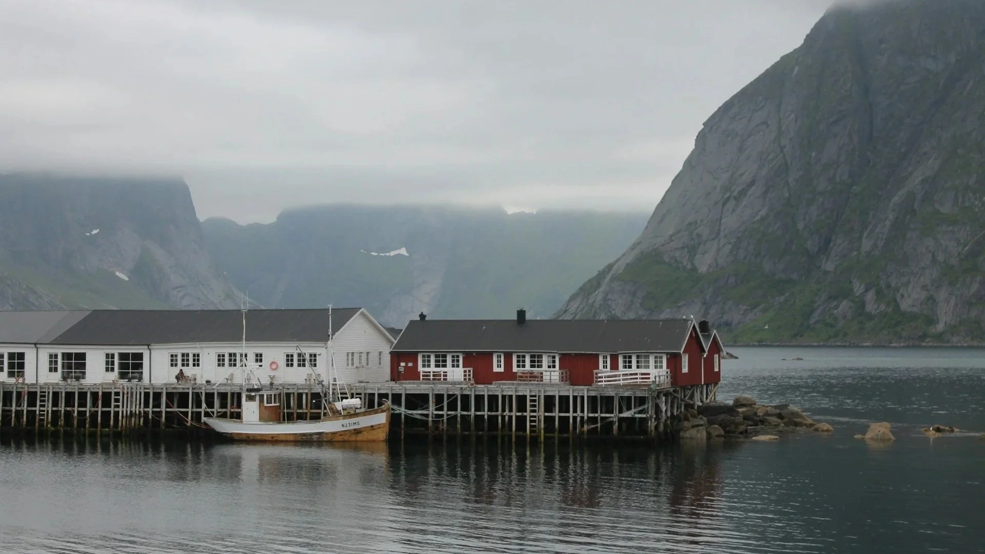 Red and white traditional fishing huts on stilts over calm water, set against a backdrop of misty, rugged mountains.