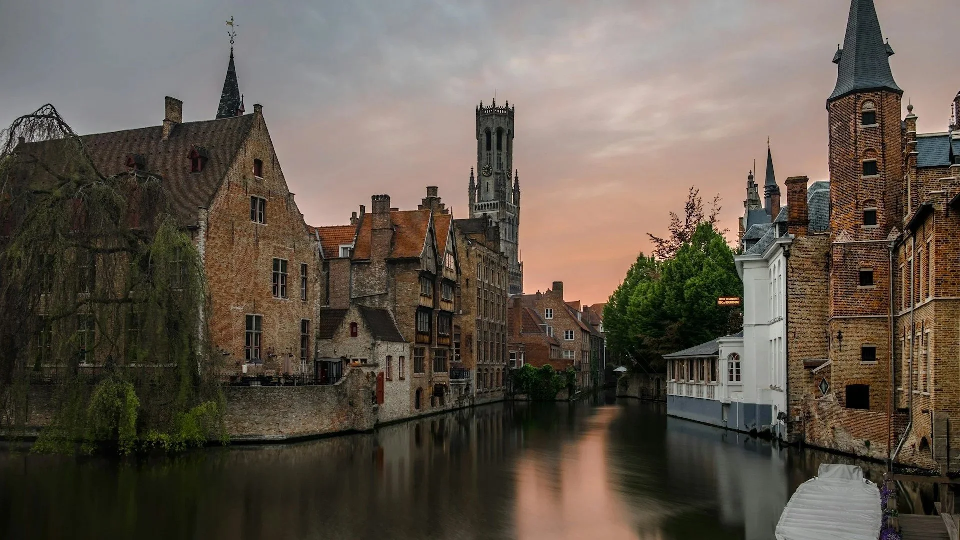 Serene canal in Bruges lined with medieval brick buildings and weeping willow trees at sunset with the Belfry tower in the distance.