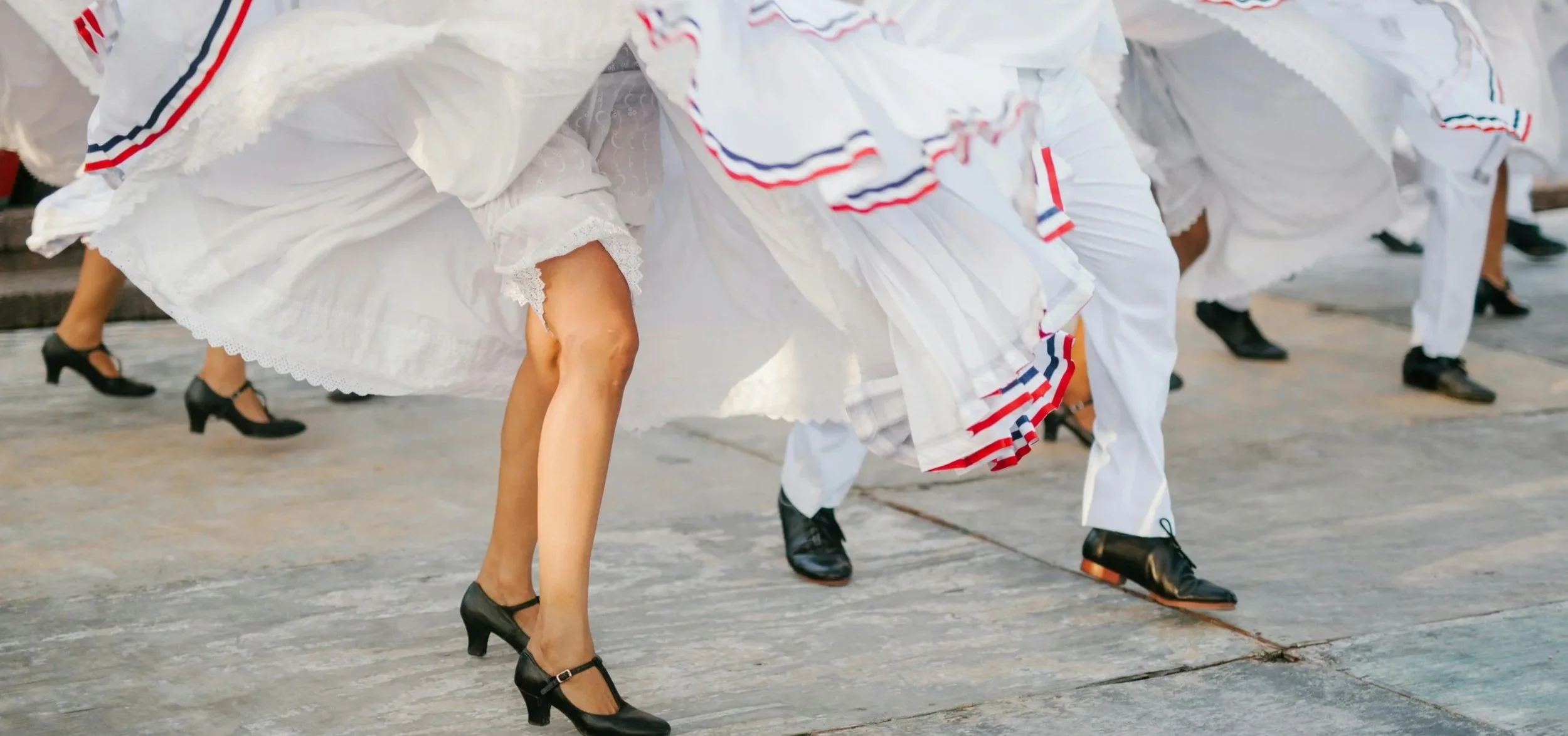 A group of dancers, dressed in white outfits with red, white, and blue ribbons, dance outdoors. They are in motion, with their skirts and pant legs swaying as they step. The image is cropped to show their legs and the lower half of their bodies.