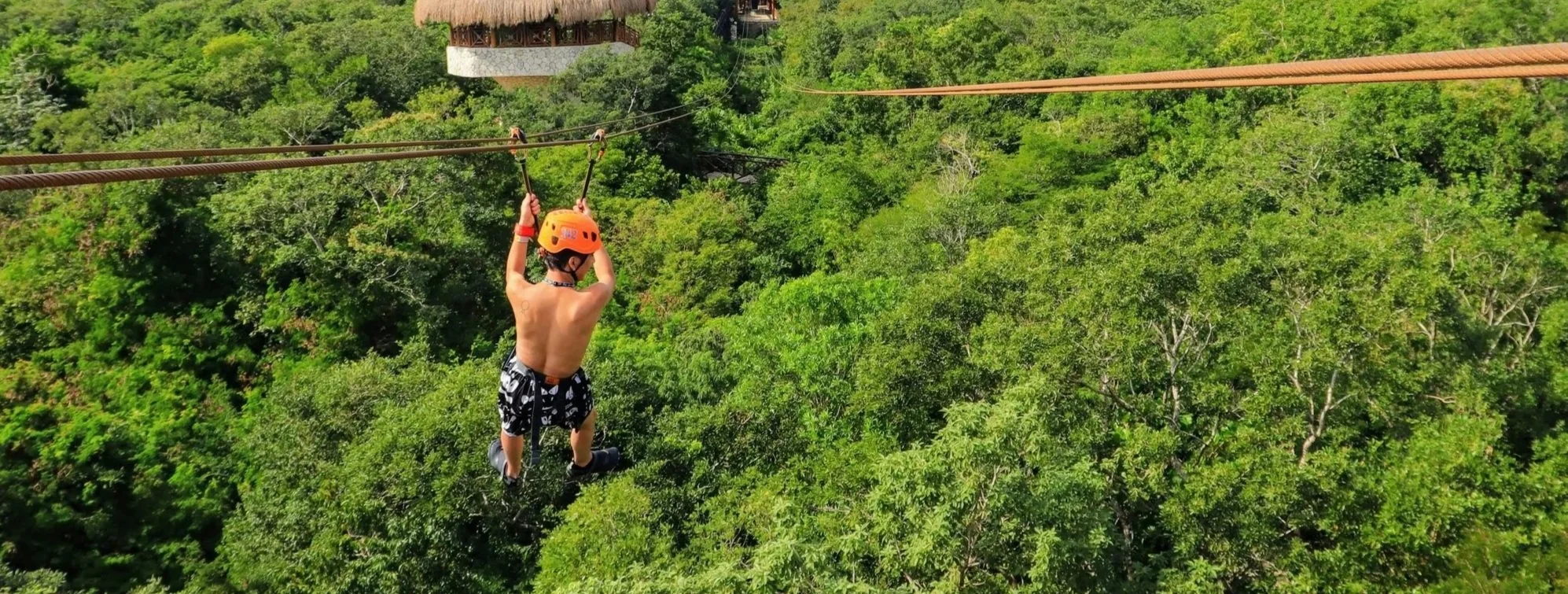 A shirtless man with an orange helmet, ziplining over a lush jungle.