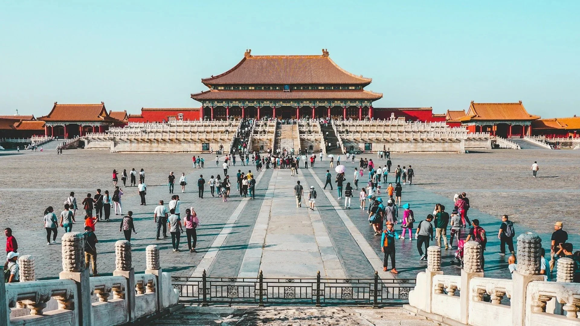 Wide view of tourists walking across the grand courtyard towards the imperial Hall of Supreme Harmony in the Forbidden City under a clear blue sky.