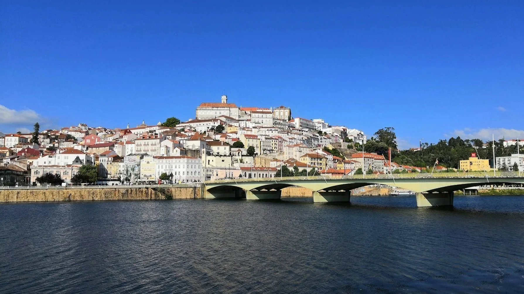 A view of the city of Coimbra rising up a hillside from the banks of the Mondego River, seen behind a wide bridge.