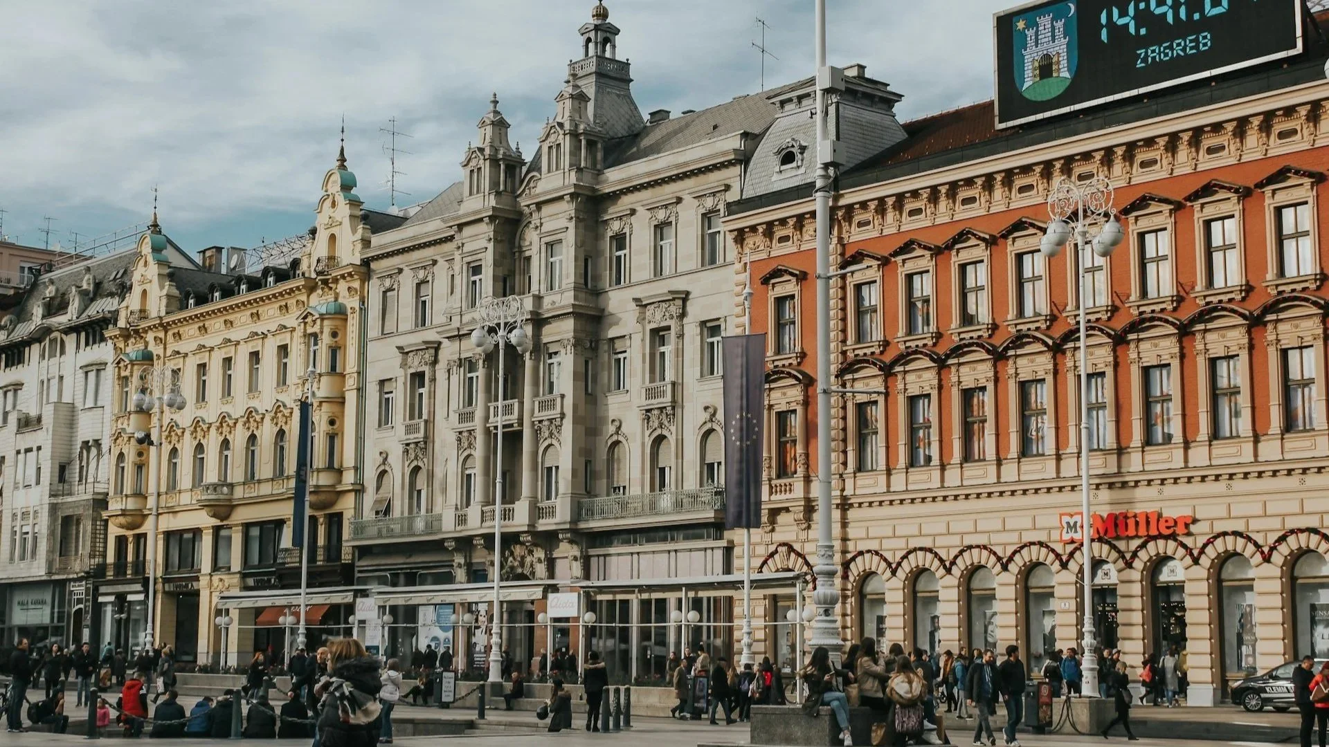 A busy city square in Zagreb, Croatia, featuring historic European architecture and a large digital clock.
