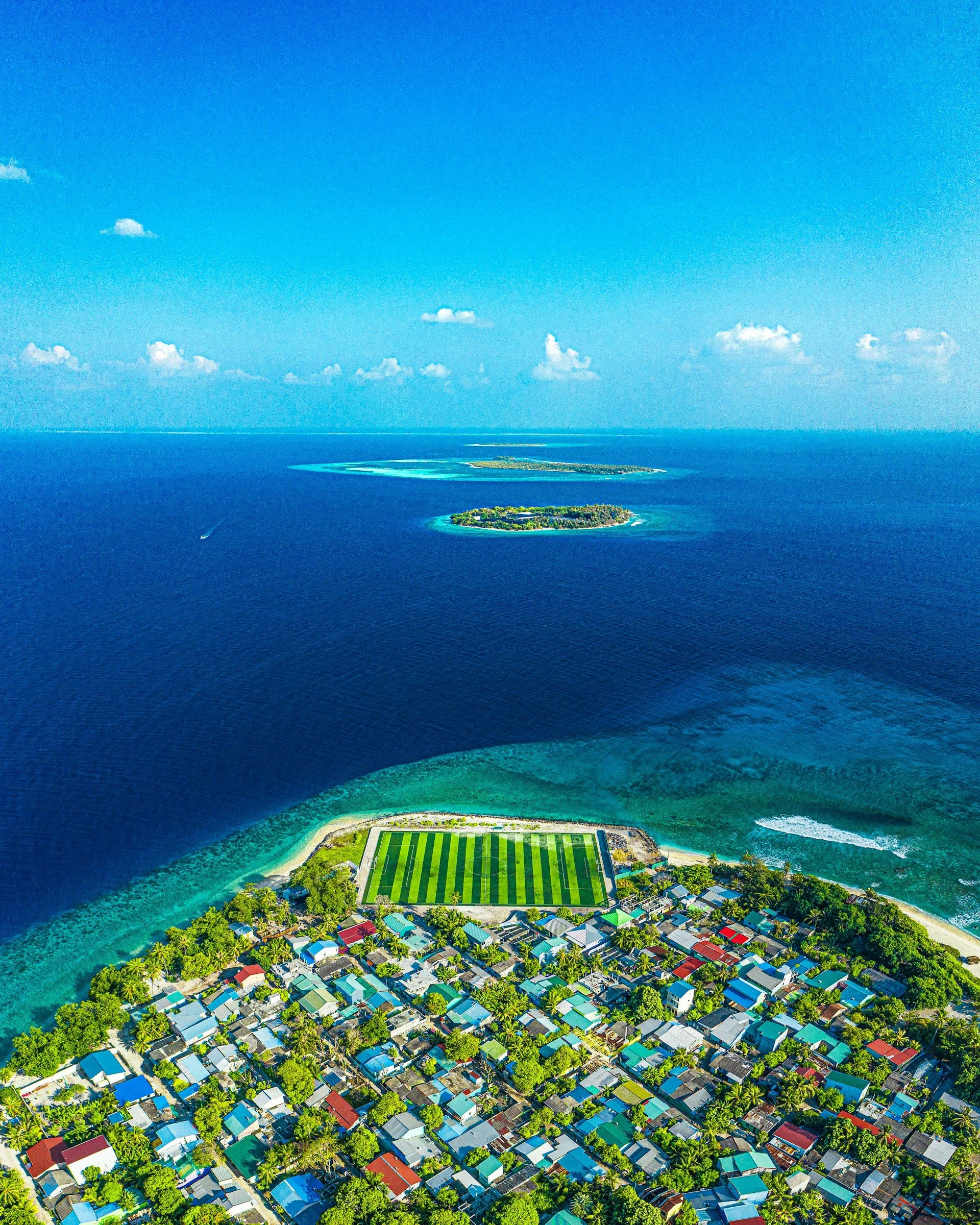 An aerial shot of a colorful coastal town on a tropical island, featuring a bright green soccer field near the turquoise shoreline.