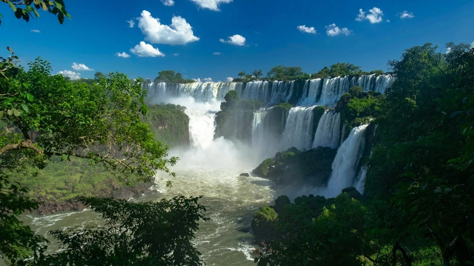 A wide, powerful waterfall cascading over a lush green cliffside under a vibrant blue sky with fluffy white clouds.