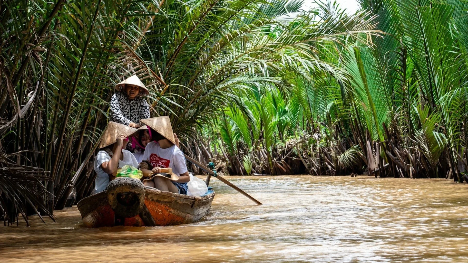 Tourists in traditional conical hats riding a small boat through a narrow, palm-lined canal.