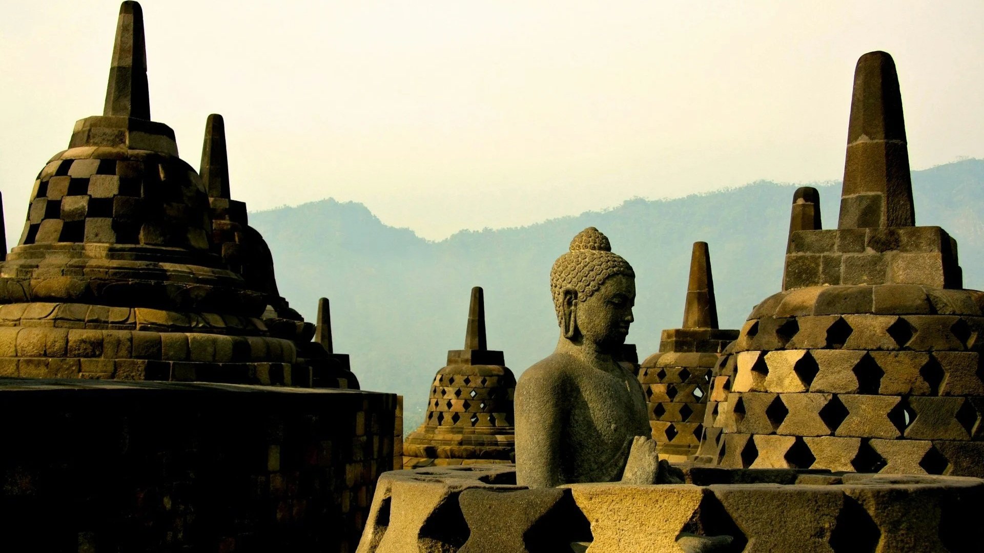 A stone Buddha statue sits serenely among several bell-shaped perforated stupas at an ancient temple during a misty sunrise.