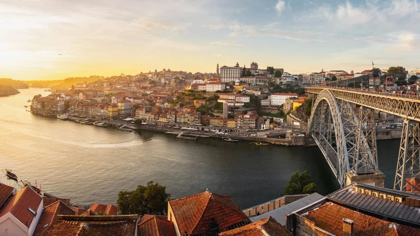 A wide panoramic view of Porto, Portugal, at sunset, featuring the Dom Luís I Bridge over the Douro River and terracotta rooftops.