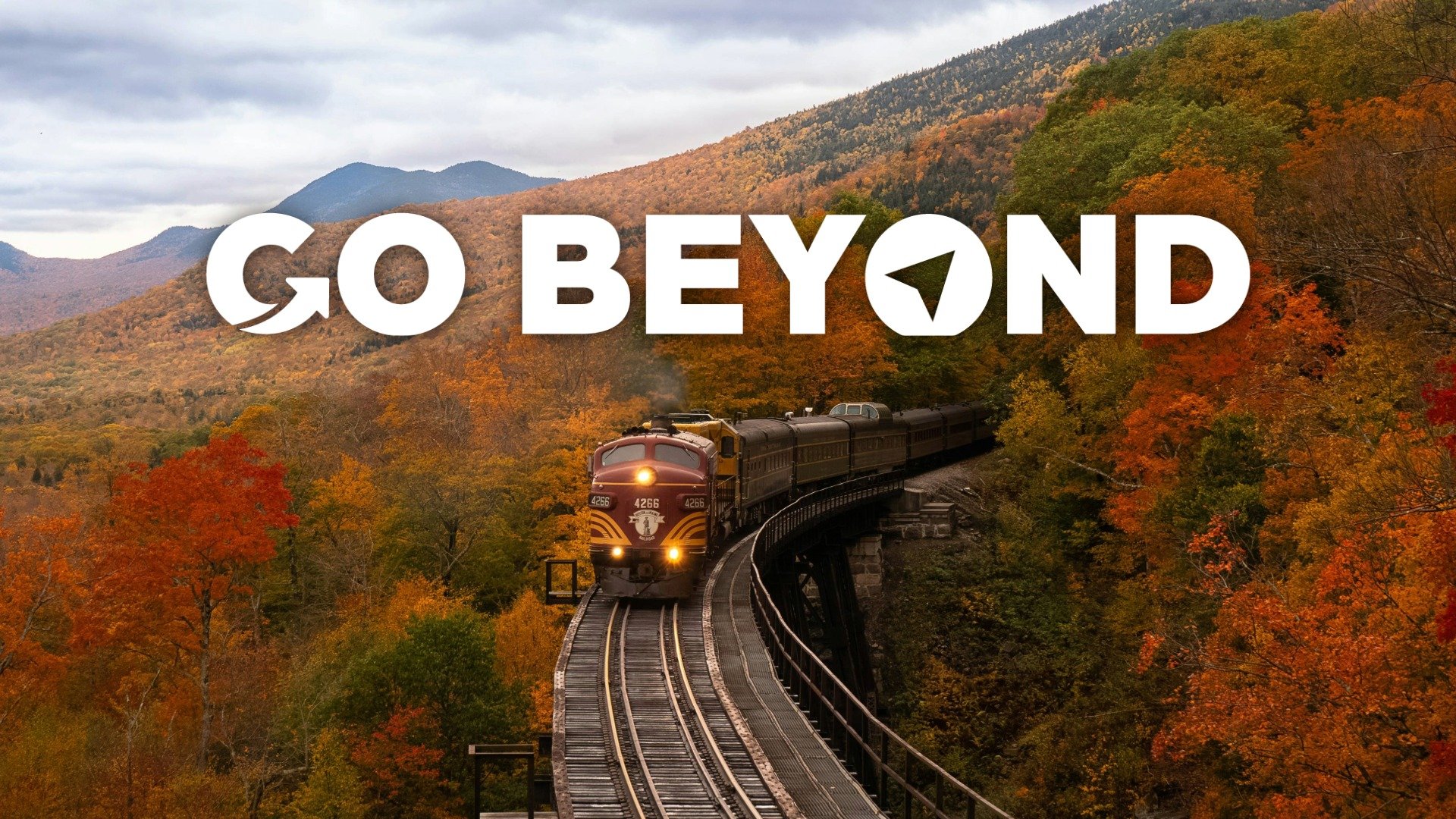 A vintage red and yellow locomotive pulling a train across a high trestle bridge through a forest of vibrant orange and yellow autumn leaves, with the "GO BEYOND" logo overlaid.