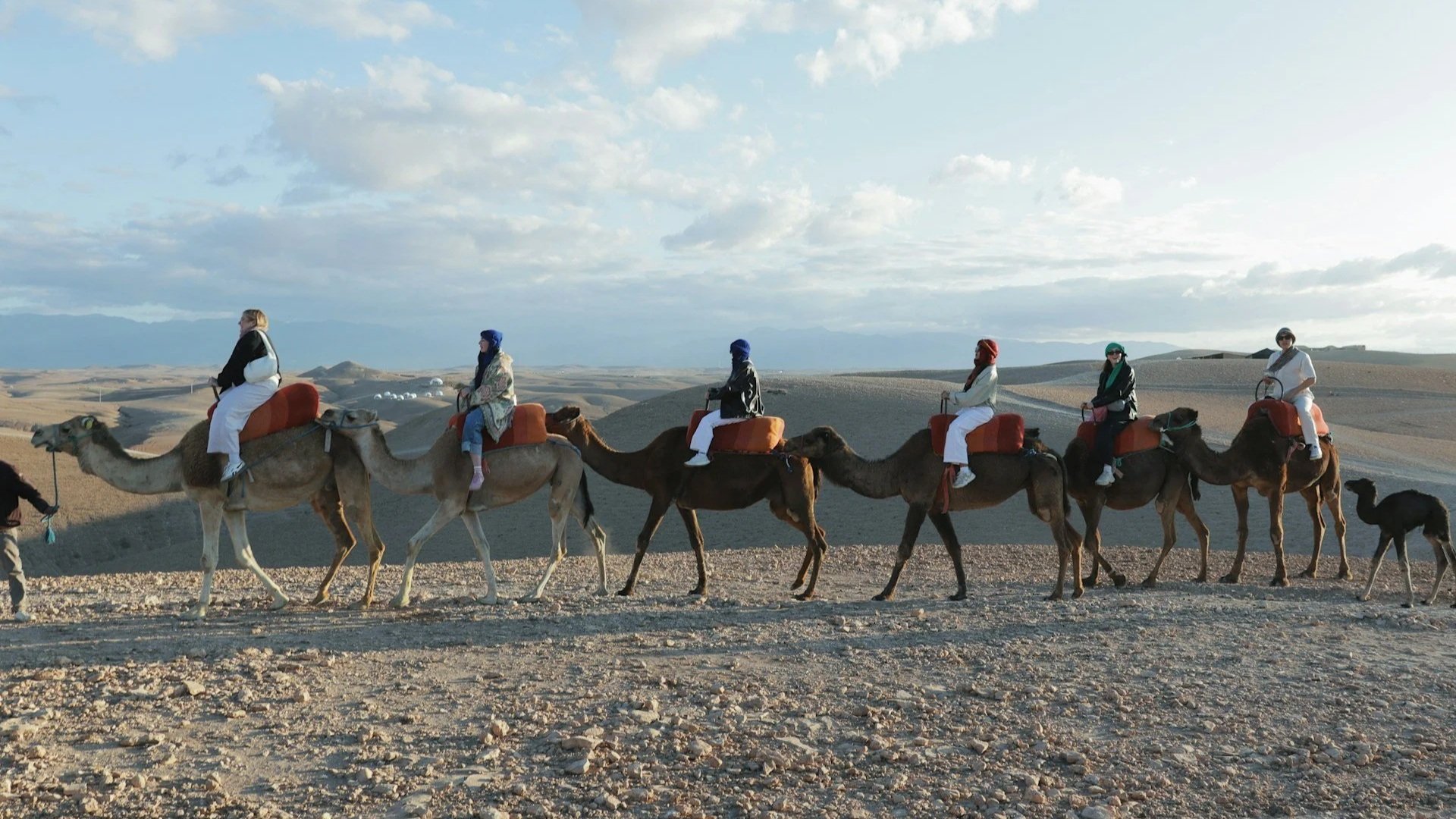 A line of travelers riding camels across a rocky desert landscape under a pale blue sky with light clouds.