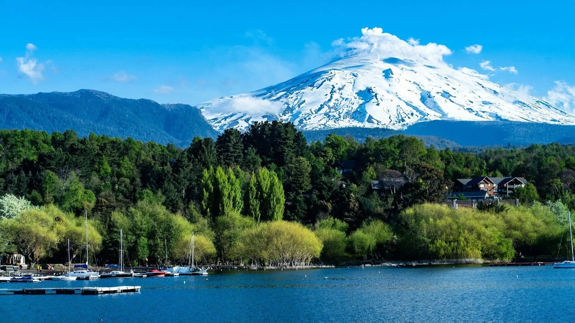 The snow-capped Villarrica Volcano looming over a lush green shoreline and a blue lake with sailboats.