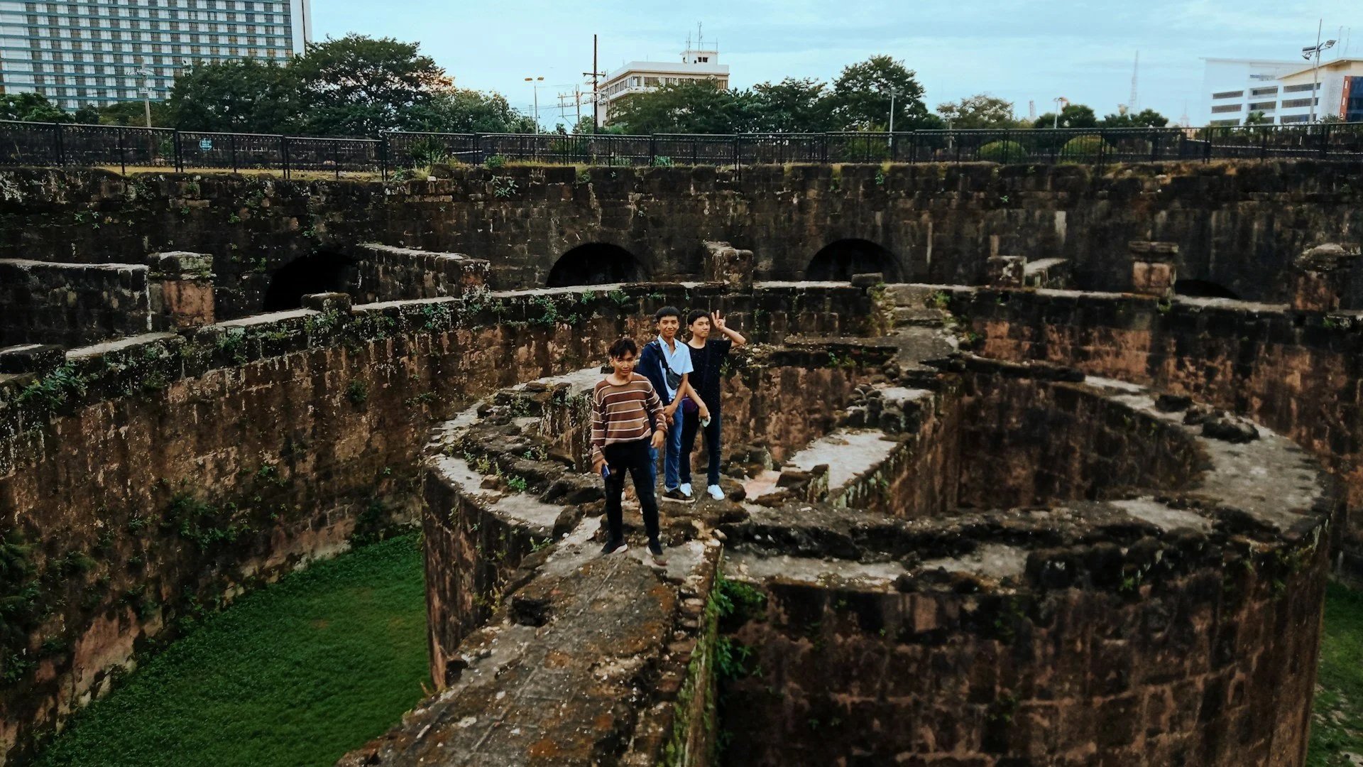 Three people standing on the circular, weathered stone ramparts of an old fort, with modern buildings visible in the background under a cloudy sky.