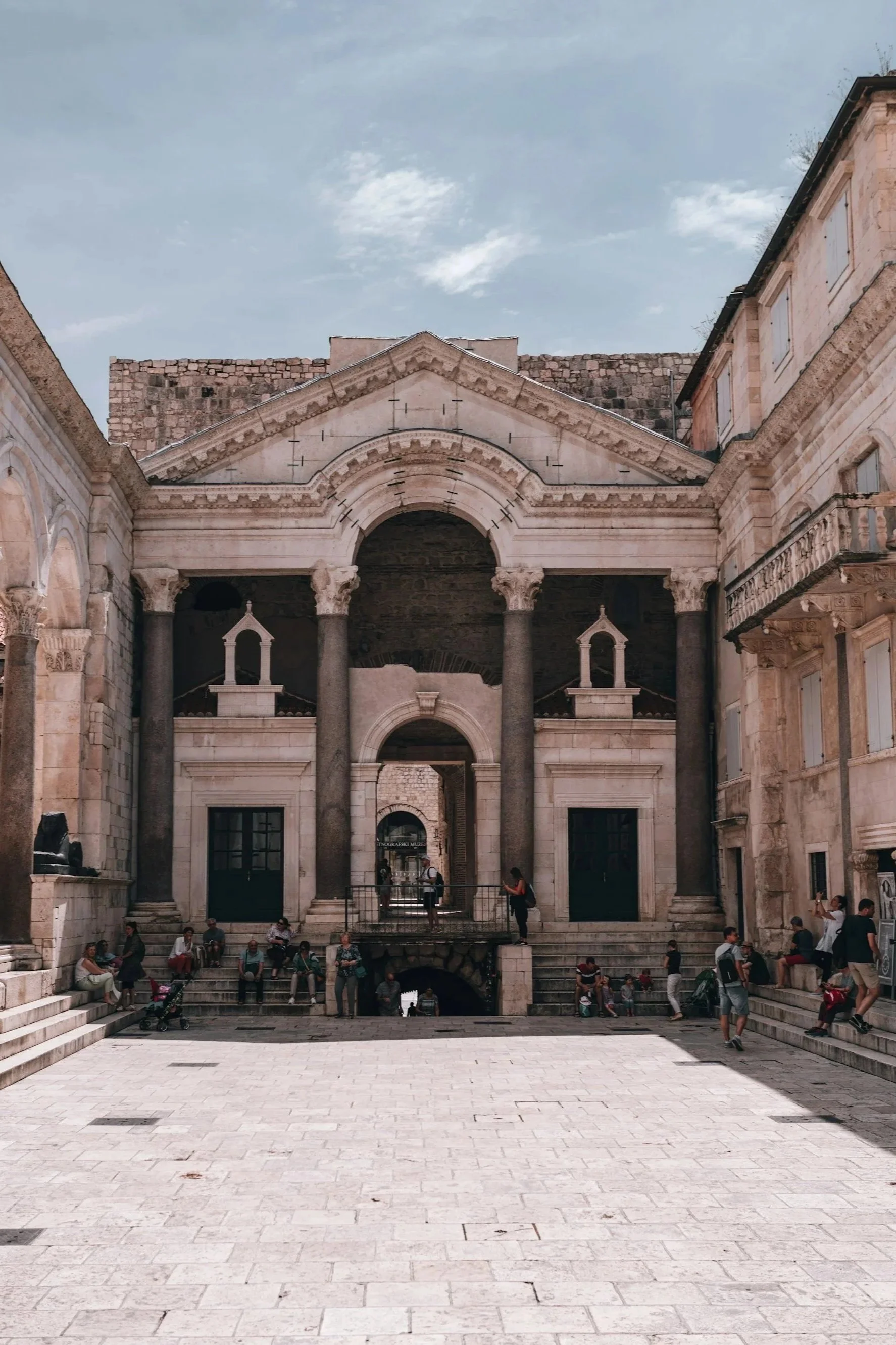 The Peristyle of Diocletian's Palace in Split, showing ancient Roman columns and arches surrounding a stone-paved courtyard with people resting on the steps.