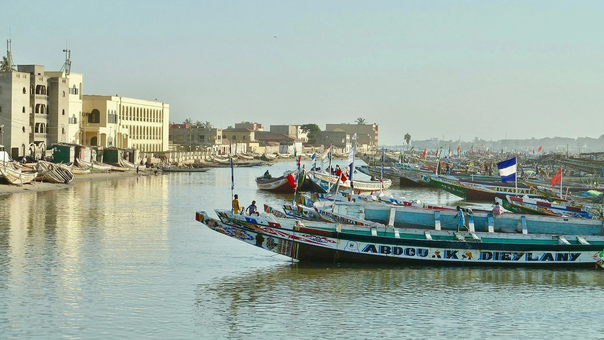 Brightly painted, traditional wooden fishing boats crowded along a calm riverbank in front of light-colored city buildings under a clear sky.