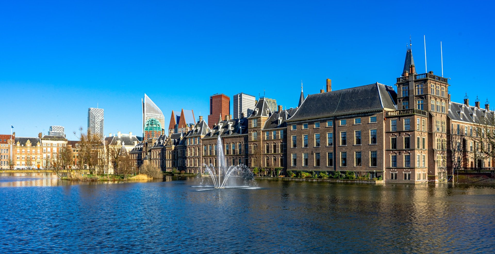 A large, historic brick building complex sits along the edge of a pond with a central fountain, with modern city skyscrapers visible in the background.