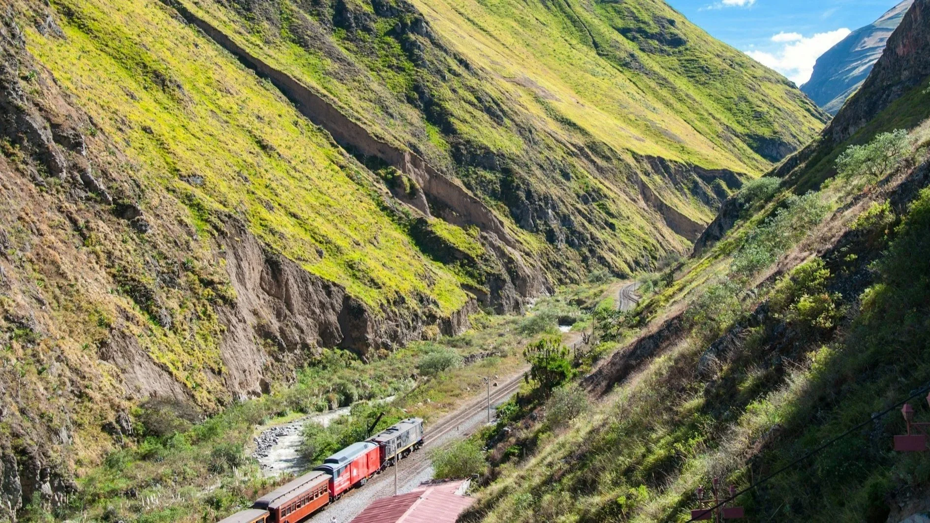 A red train travels along a narrow valley floor between steep, grassy mountain slopes under a bright blue sky.