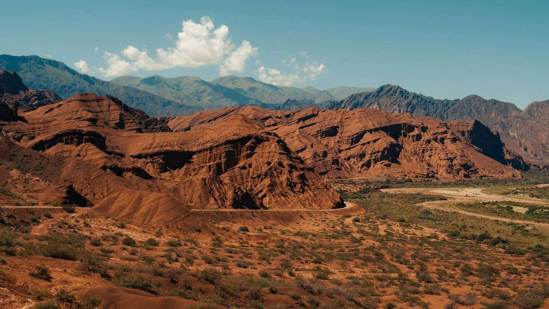 A wide landscape of rugged, reddish-brown rock formations and canyons under a clear blue sky with sparse green shrubs in the foreground.