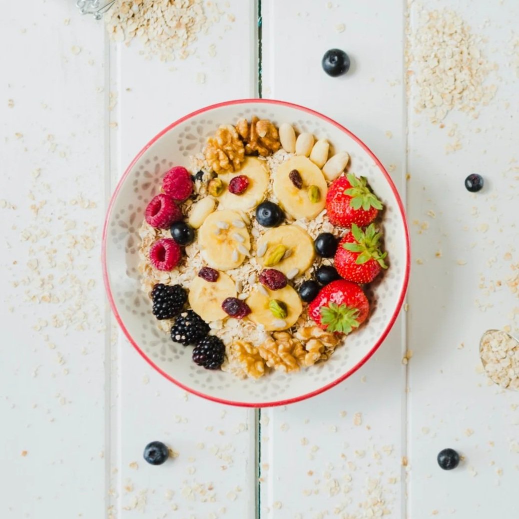 A top-down view of a white wooden table featuring a bowl of Bircher muesli topped with bananas, strawberries, berries, and walnuts, surrounded by scattered oats and a small glass jar.
