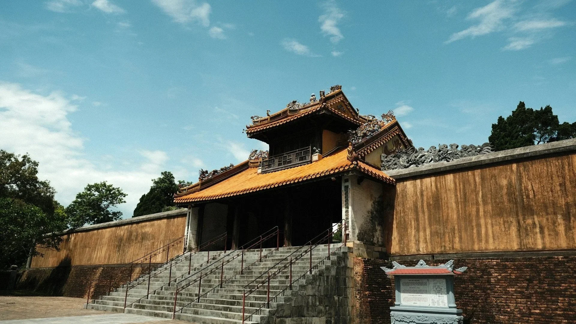 A wide shot of a weathered stone gate with a traditional tiered yellow roof and dragon carvings, set against a long masonry wall and a bright blue sky with wispy clouds.