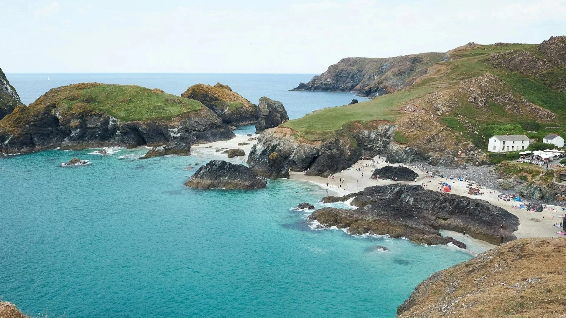 A scenic view of Kynance Cove in Cornwall, featuring turquoise water, rocky islands with green tops, and a small sandy beach with people.