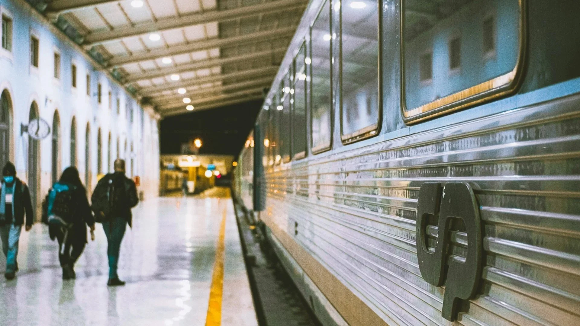A stainless steel CP (Comboios de Portugal) train stopped at a modern station platform at night with passengers walking alongside.
