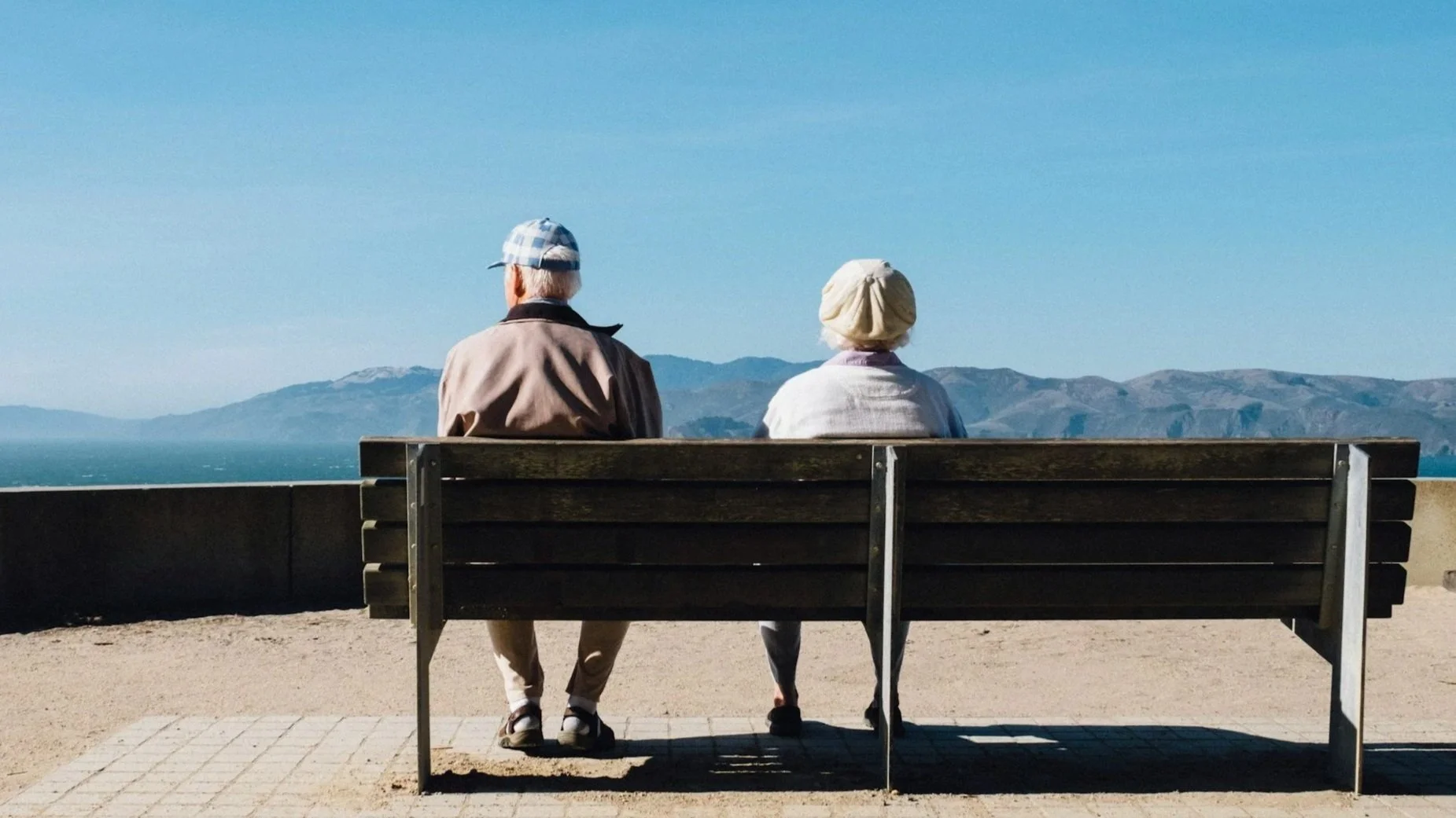 Rear view of an elderly couple sitting on a wooden bench, looking out at a calm sea and distant mountains.