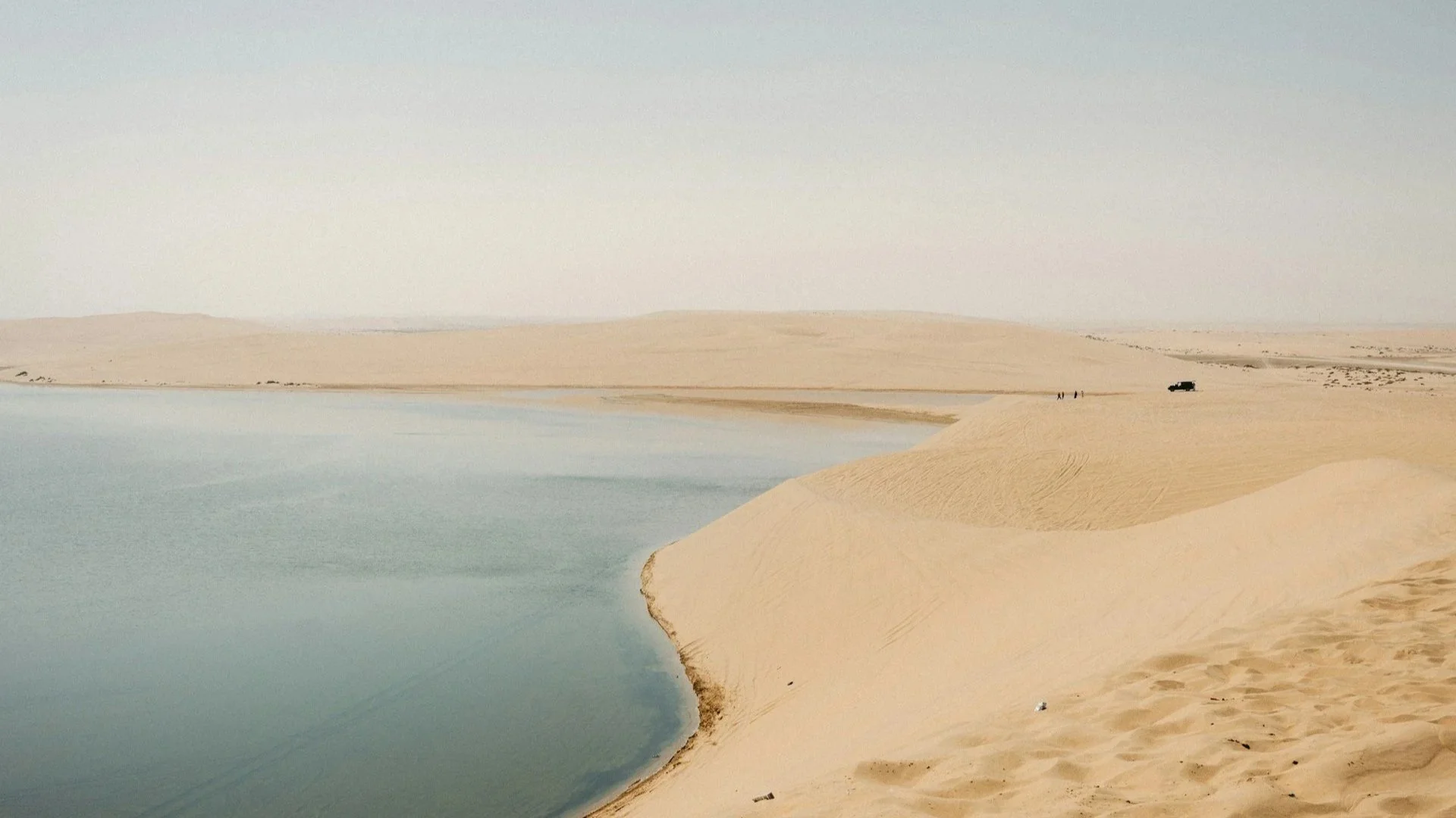 A vast, rolling sand dune meet the calm blue waters of the Inland Sea in Qatar under a clear sky.