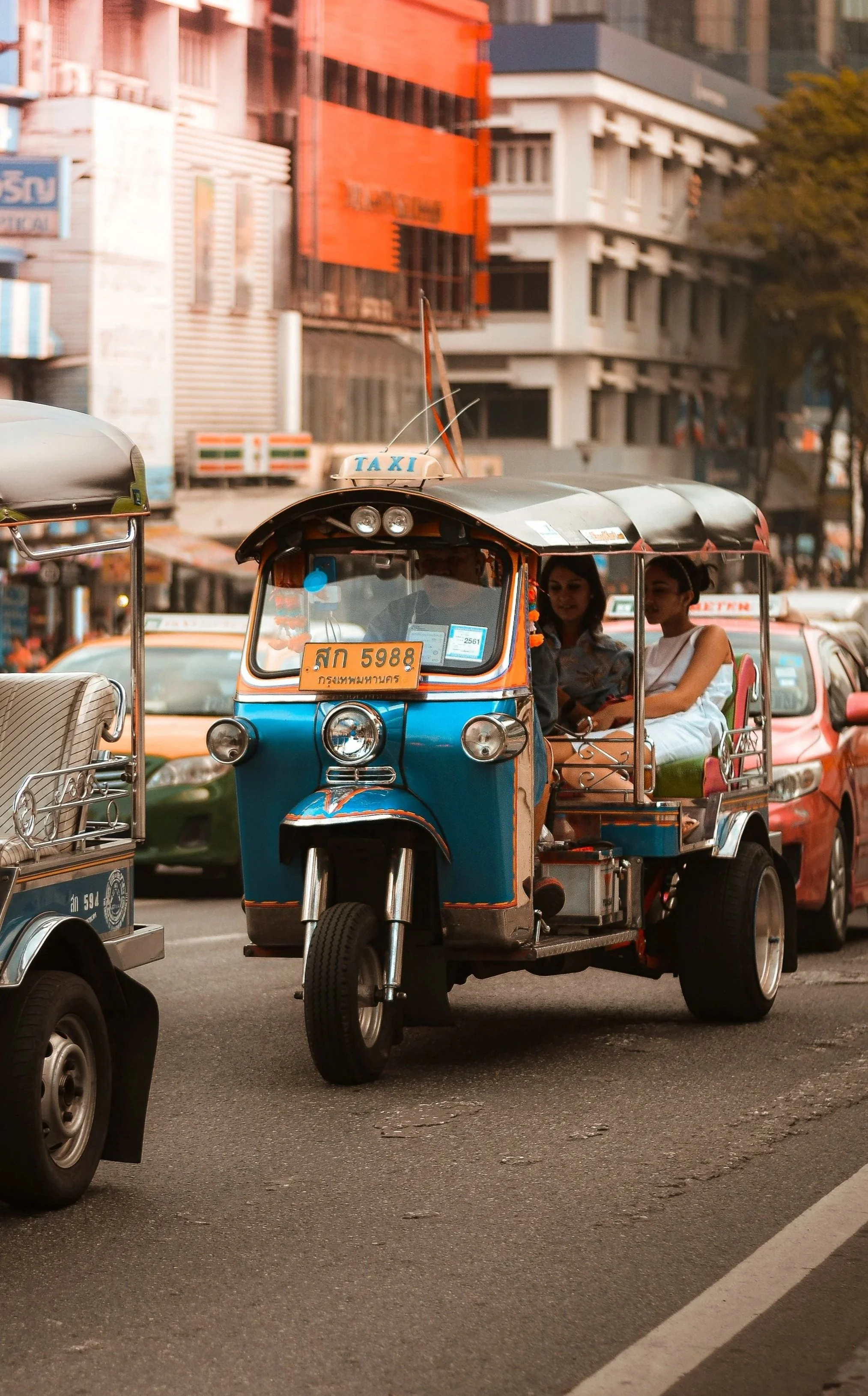 A blue and red tuk-tuk taxi on a busy street in an urban area of Thailand.