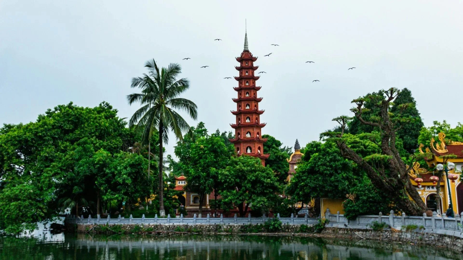 The tall, red Tran Quoc Pagoda surrounded by lush green trees next to West Lake in Hanoi.