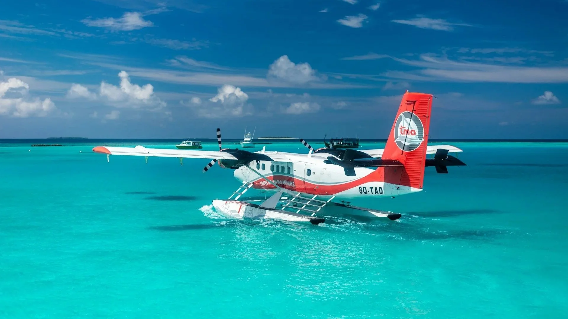A red and white seaplane floats on vibrant turquoise water under a blue sky with scattered clouds.