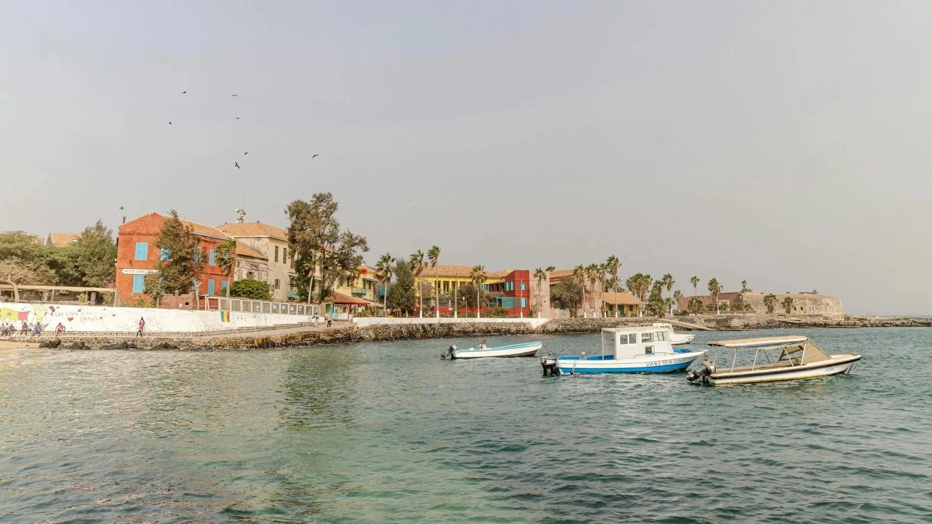 Several small motorboats floating on calm coastal water near a shoreline lined with colorful colonial-style buildings and palm trees.