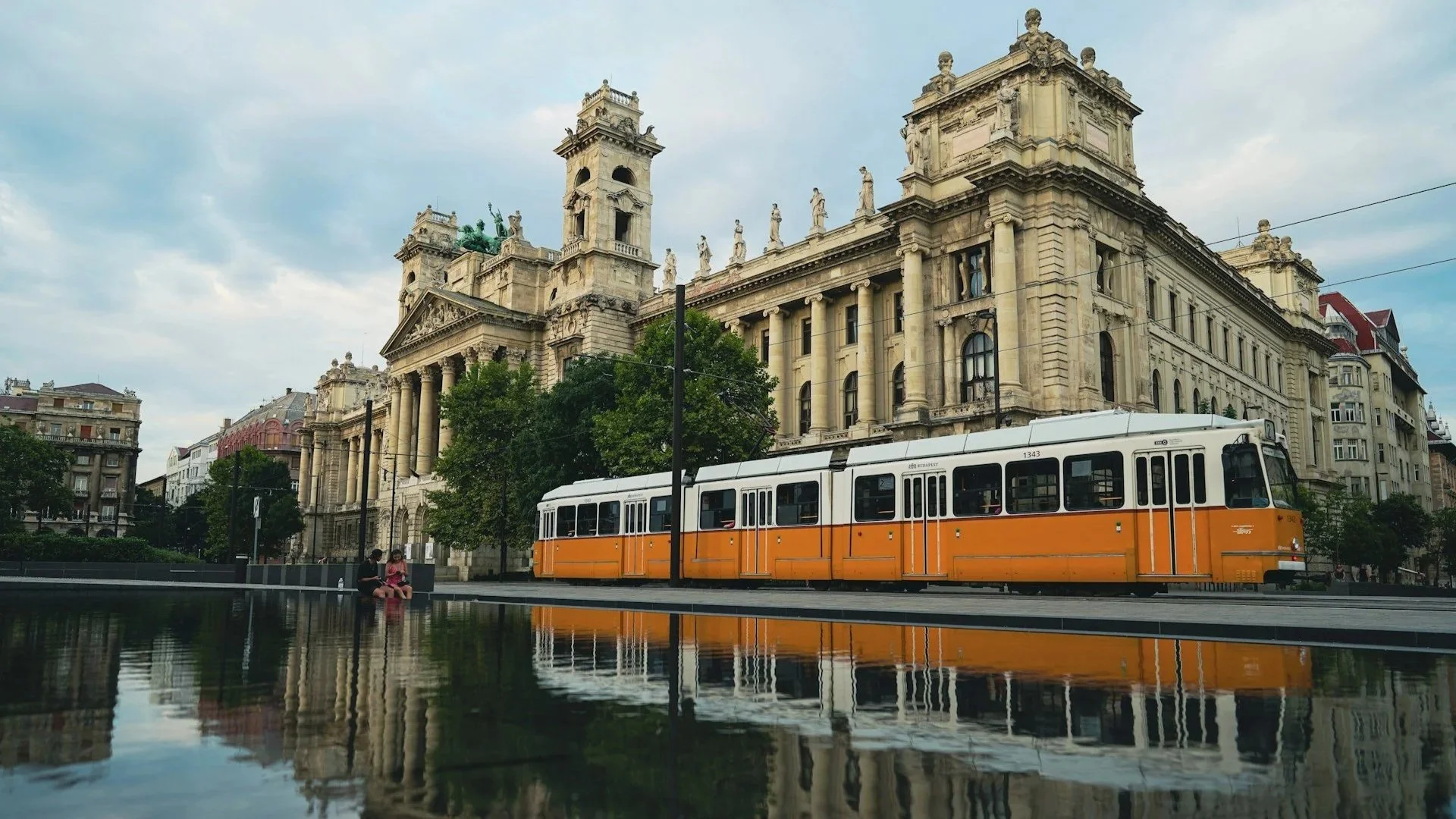 Yellow tram driving past a grand classical building in Budapest, reflected in a still pool of water.