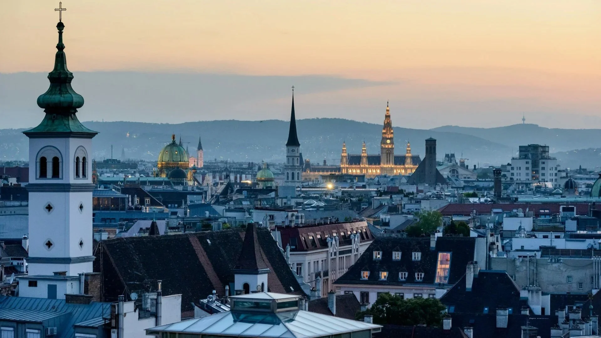 A twilight city view of Vienna, highlighting various historical spires and the illuminated Rathaus (City Hall) under a soft evening sky.