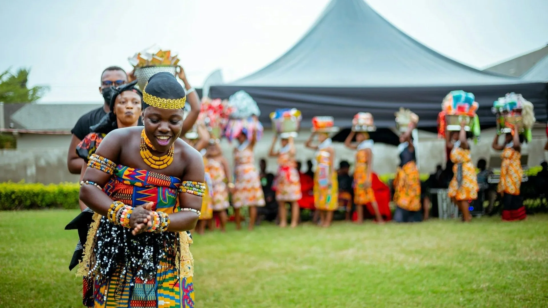 A woman in traditional Ghanaian kente cloth and beaded jewelry dances outdoors, followed by a line of people carrying gift baskets on their heads.