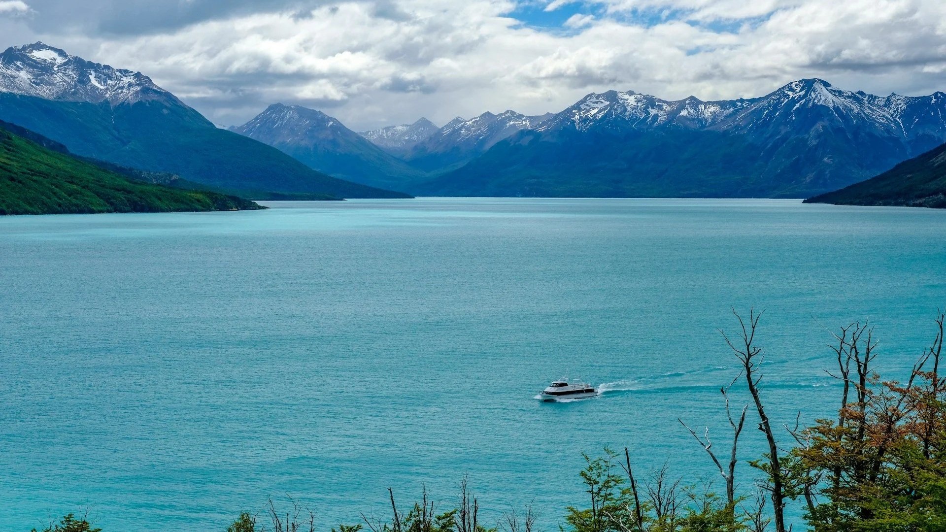 A white tour boat cruising across a large, vibrant turquoise lake surrounded by snow-capped mountains under a cloudy sky.