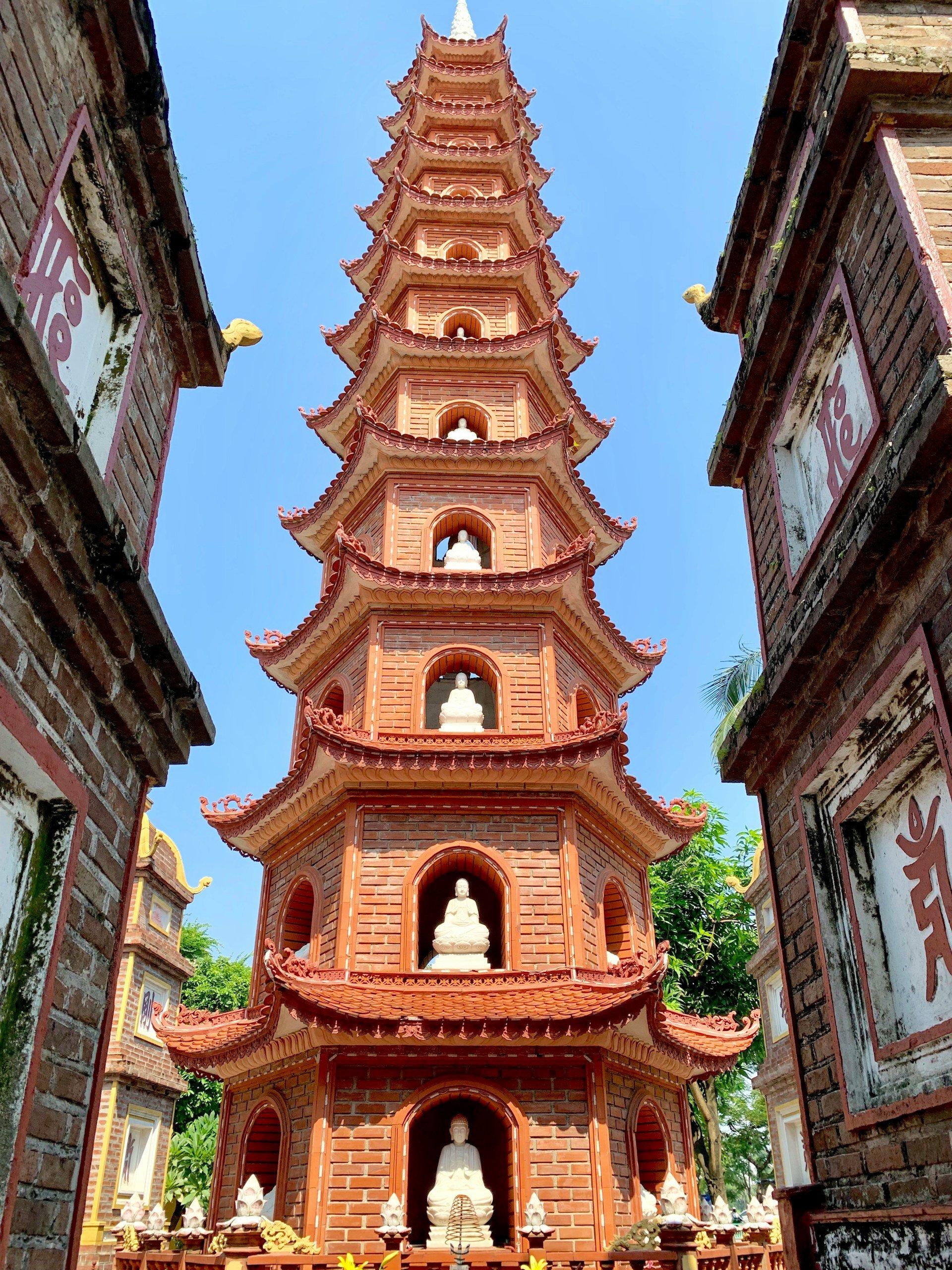 A low-angle shot of the towering, eleven-story red brick Tran Quoc Pagoda in Hanoi, featuring white Buddha statues nestled in arched alcoves on each level.