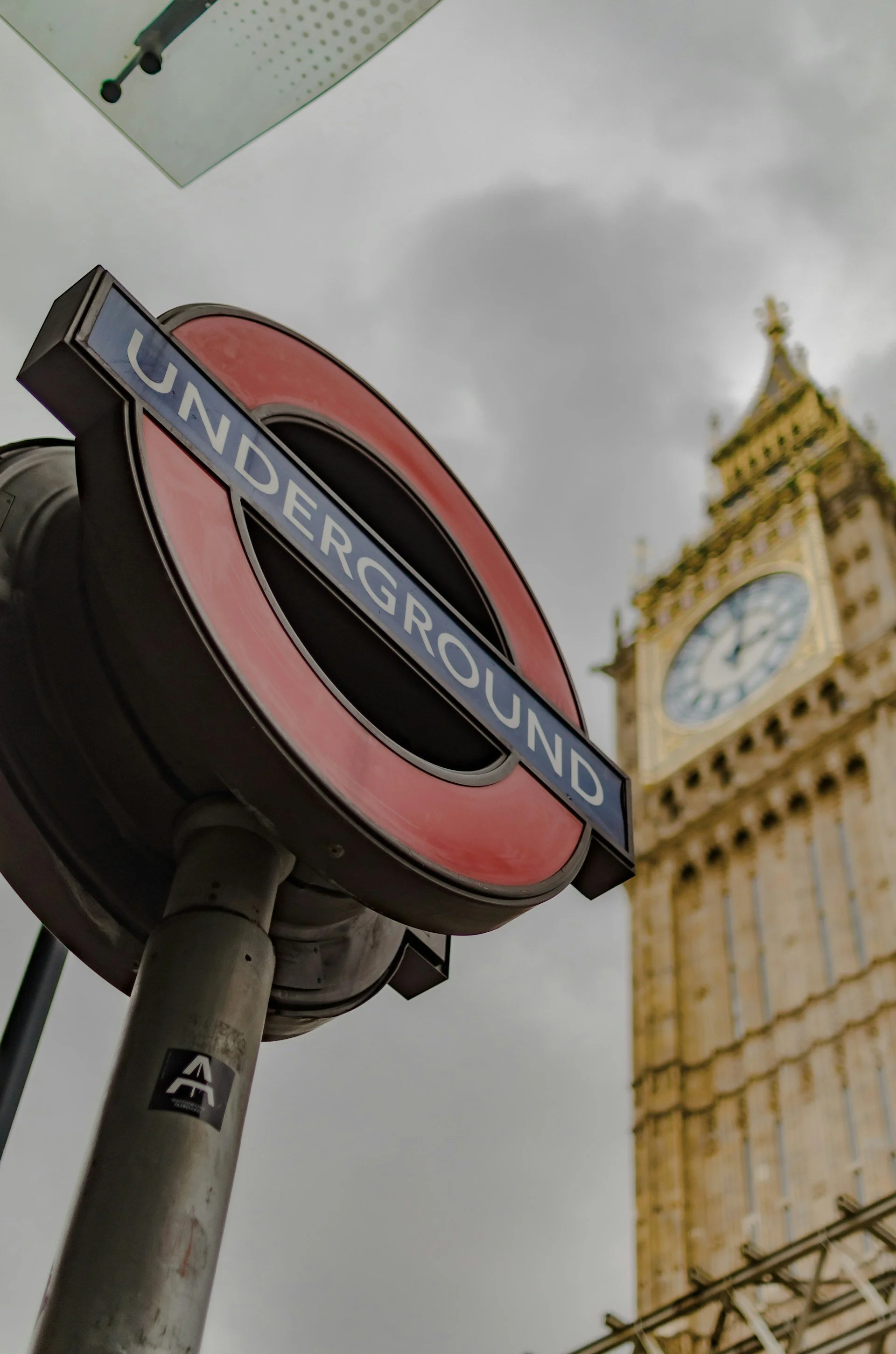 A low-angle shot of a classic red and blue London Underground roundel sign. In the soft-focus background, the iconic Big Ben clock tower rises against a moody, overcast sky.