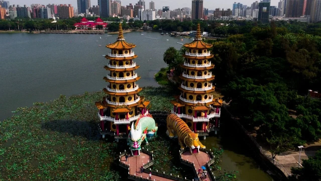 An aerial view of the Dragon and Tiger Pagodas on Lotus Pond, showcasing the colorful dragon and tiger sculptures at the entrance, surrounded by green lily pads with a modern city skyline in the background.