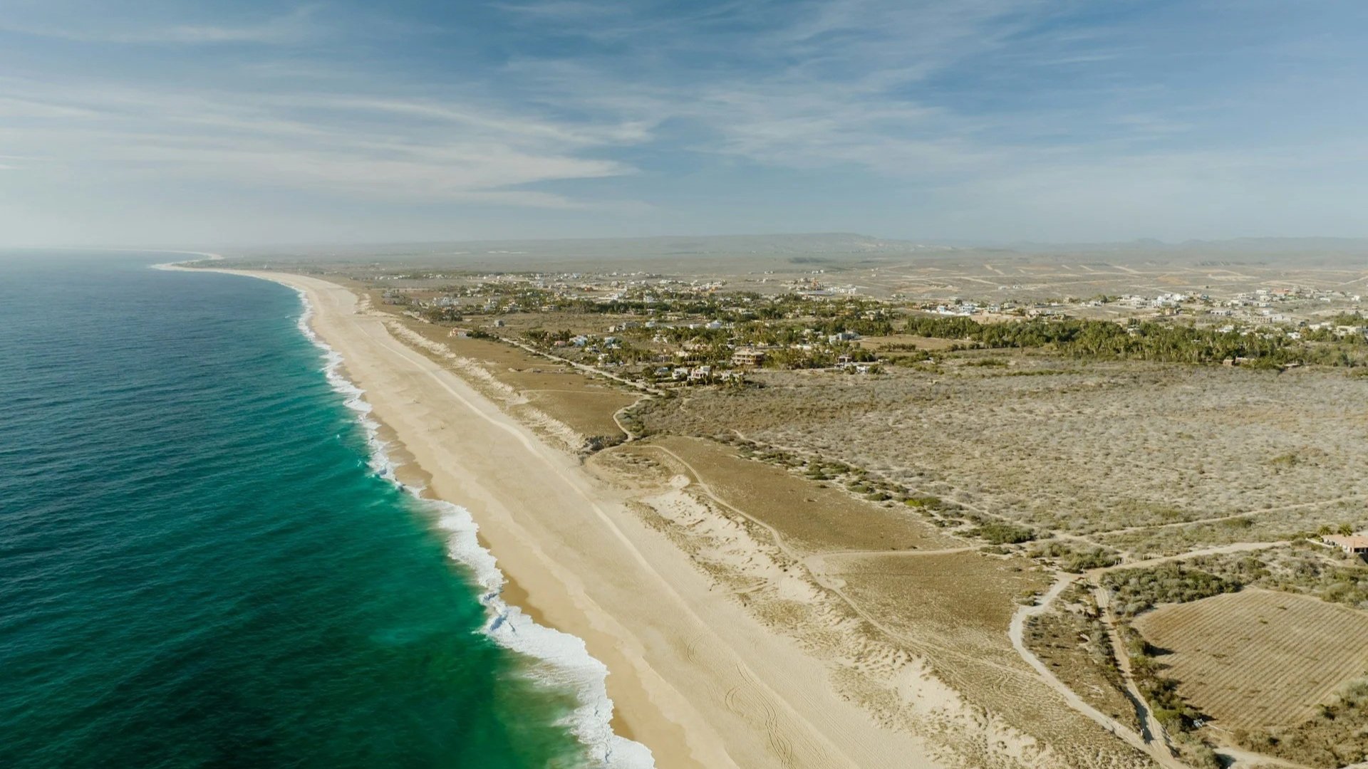An aerial view of a wide, sandy beach meeting bright turquoise ocean water, with a small coastal town and dry brushland stretching toward the horizon.